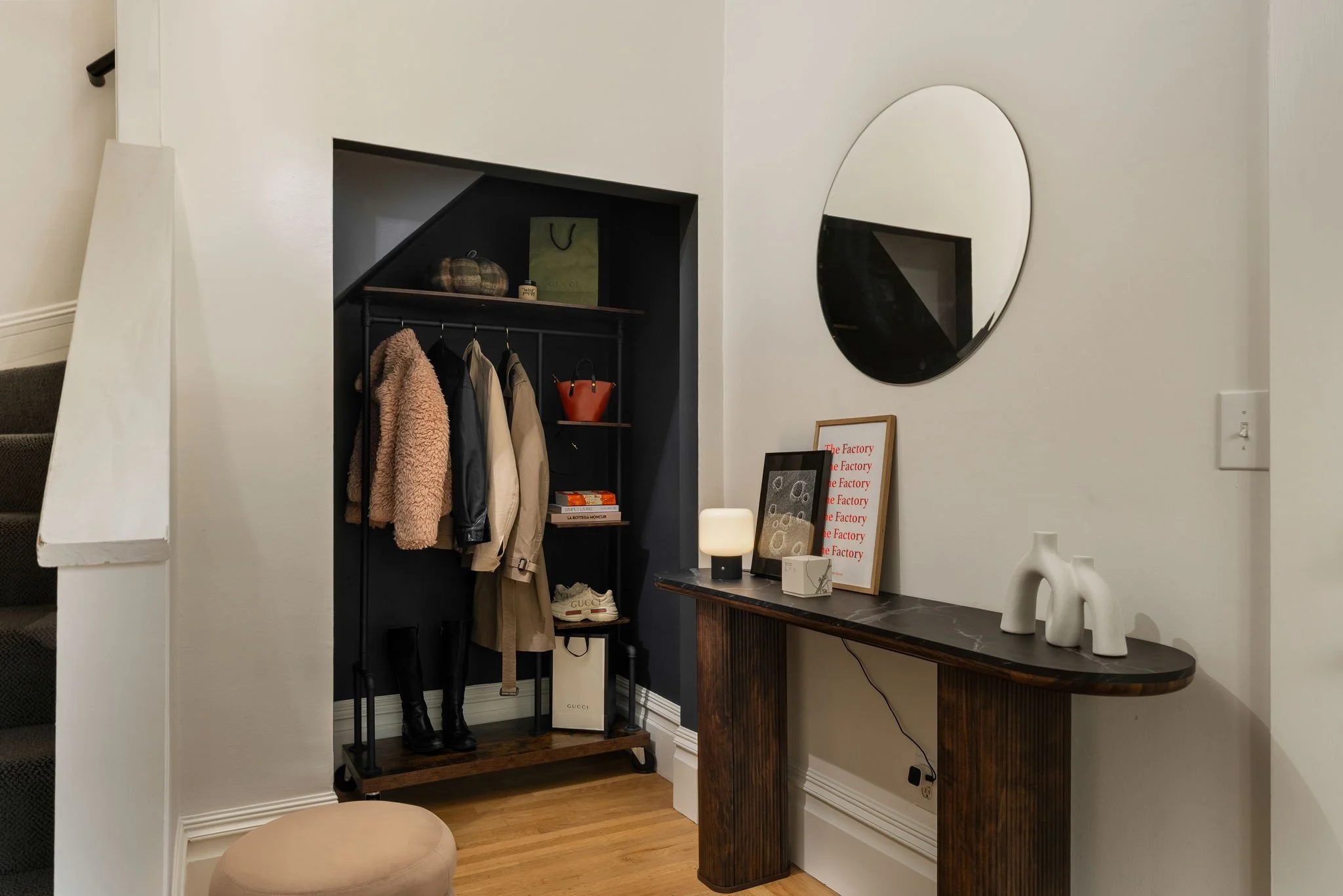 A home entryway with a black clothing rack holding jackets, a Gucci shopping bag, and a pair of white sneakers. Above the rack is a shelf with a plaid bag and a Gucci shopping bag. To the right, a small table with decorative objects including a white