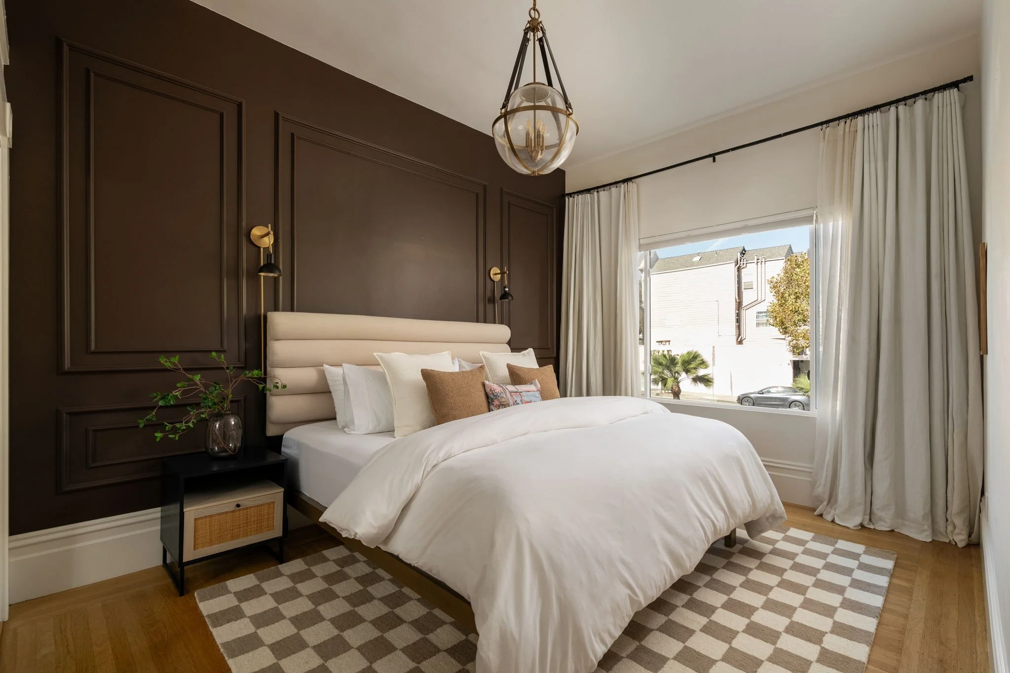 A bedroom with a large window, beige and dark brown walls, a bed with white bedding and pillows, and a small black nightstand with a glass vase and a plant. There are white curtains over the window and a pendant light hanging from the ceiling.
