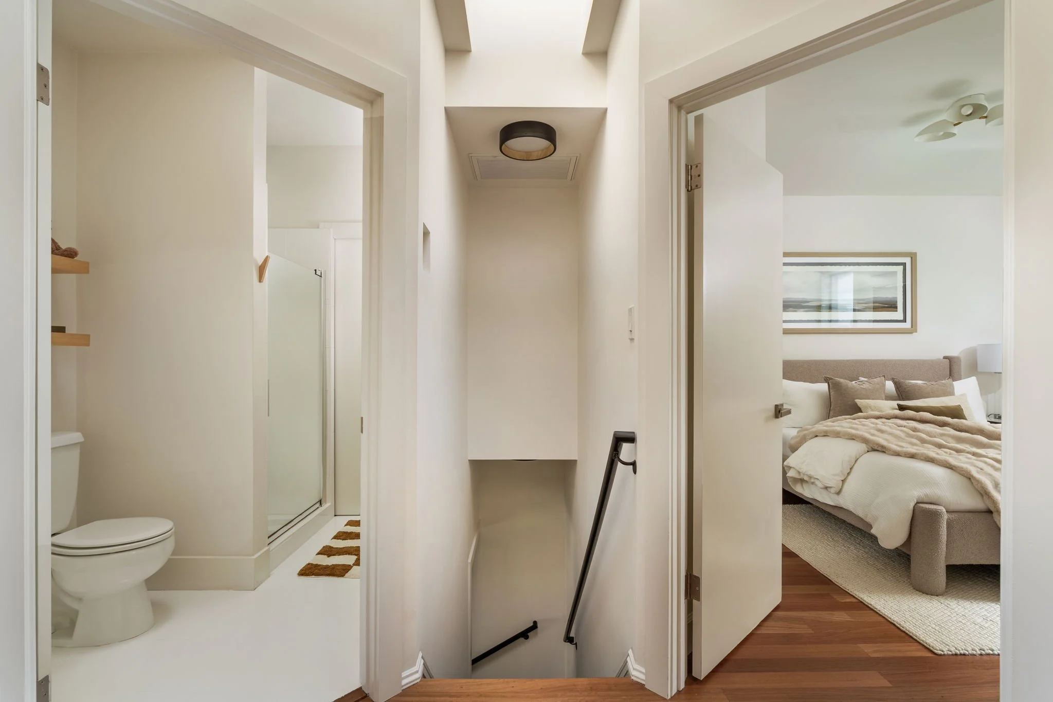 View of a small bathroom with a toilet, large mirror, and shower with glass door seen from a hallway, with a bedroom visible through an open door on the right.