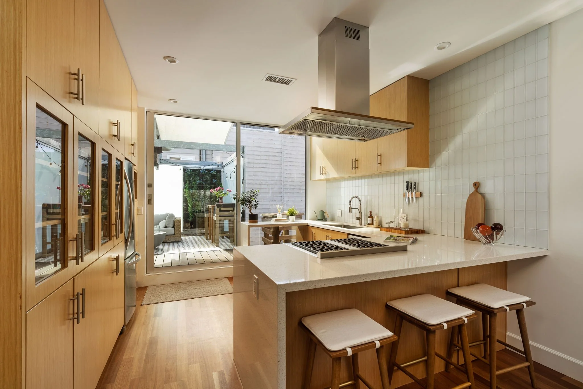 Modern kitchen with wooden cabinets, white countertops, and a breakfast bar with stools. There is a glass door leading to an outdoor patio with seating and a dining area. The kitchen has a stainless steel range hood and a tiled backsplash.