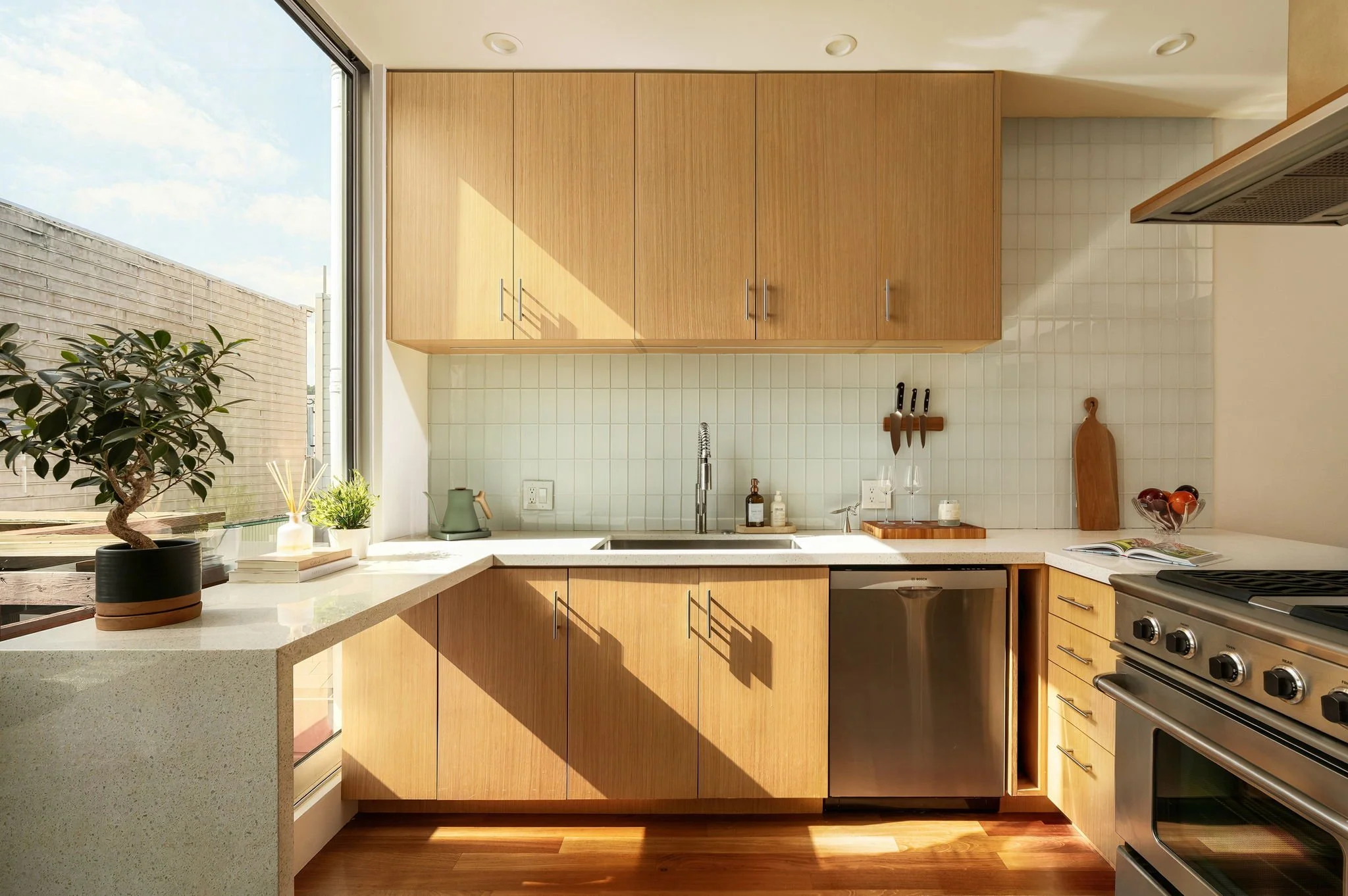 Modern kitchen with light wood cabinets, granite countertop, white tile backsplash, large window, potted plant, cutting board, and stainless steel appliances.