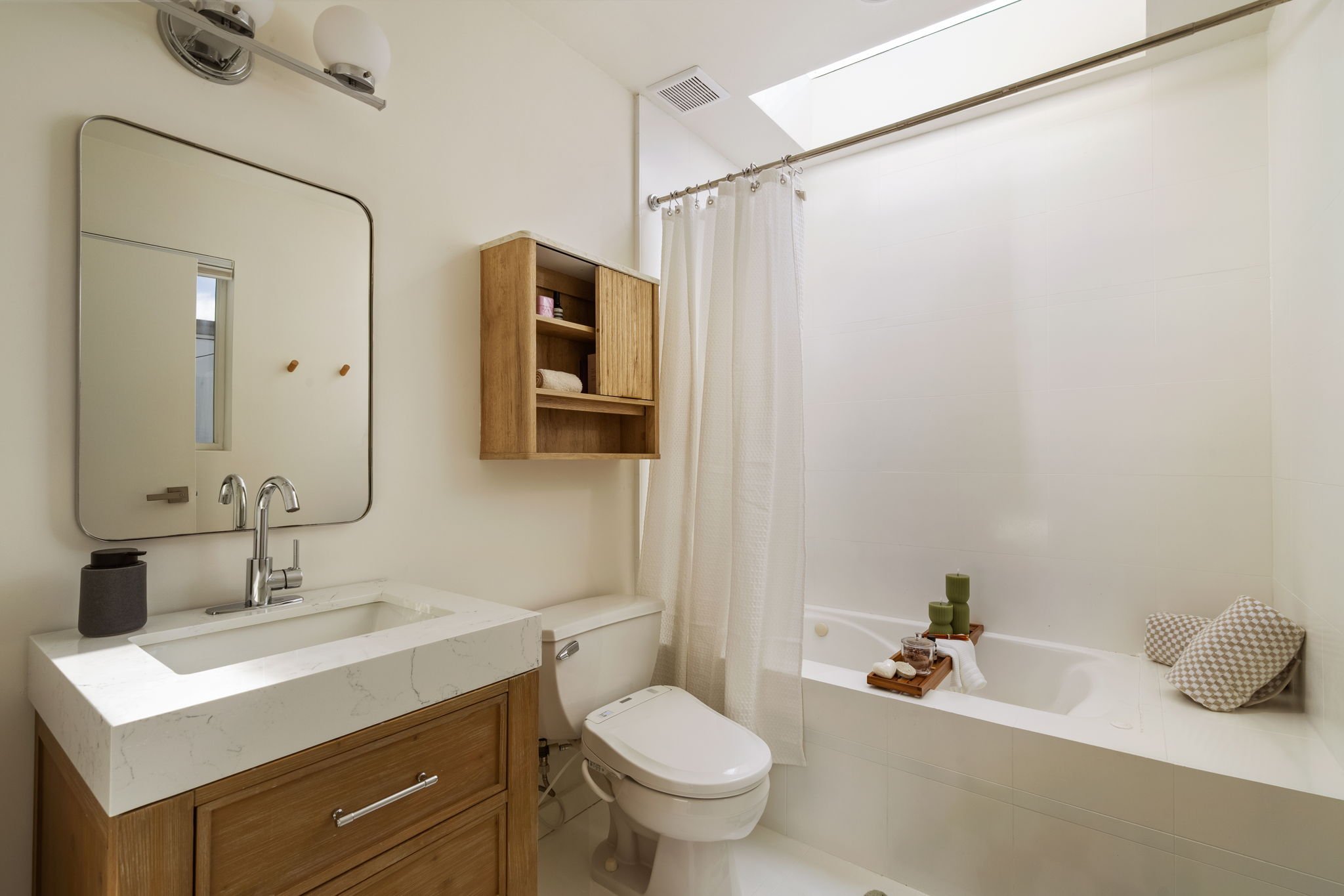 A bathroom with a wooden vanity with a white marble countertop, a rectangular mirror, a sink, a towel holder, and a faucet. There is a small window, a wooden shelf with toiletries, a shower curtain, and a bathtub. The bathroom has a skylight and mini