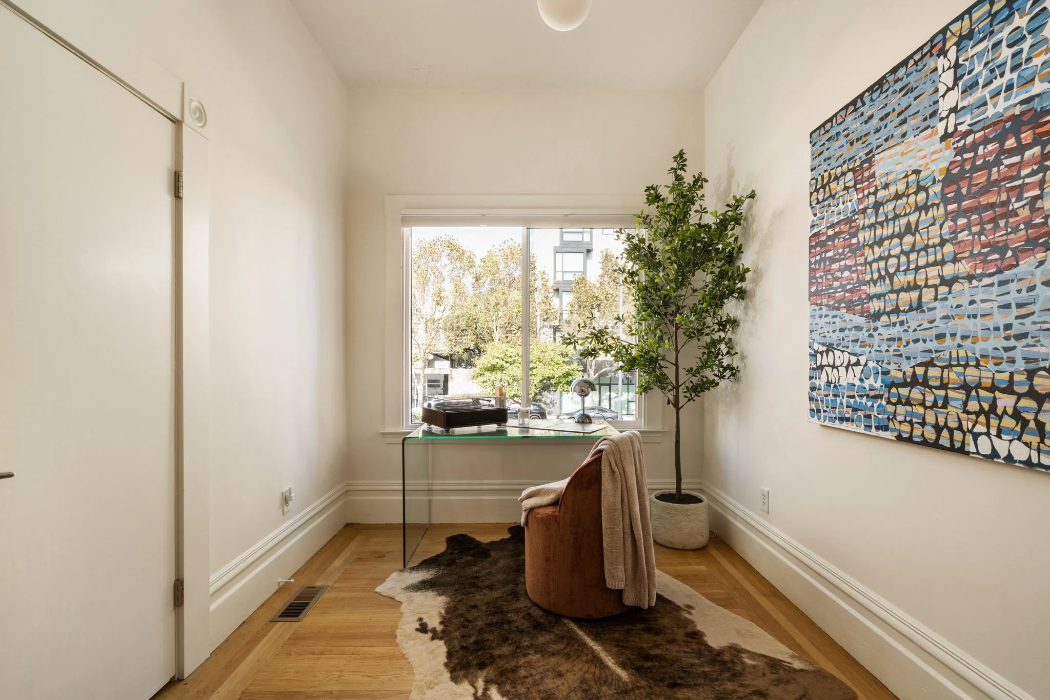 A cozy corner of a room with a large window showing trees outside, decorated with a tall indoor plant, an abstract wall art, a glass table with a turntable, a chair with a beige throw, and a cowhide rug on a hardwood floor.