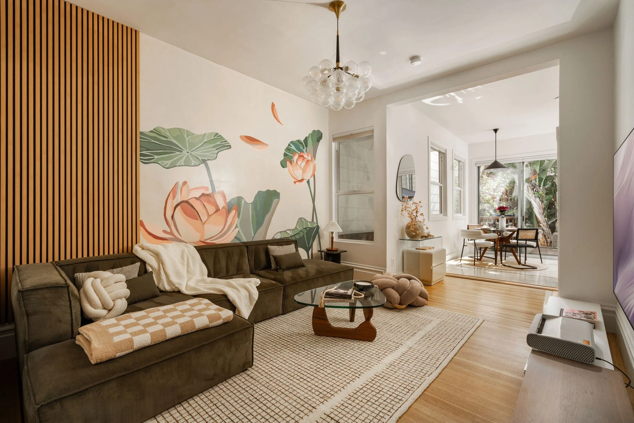 A living room with a dark gray sectional sofa decorated with throw pillows and a blanket, a modern wooden coffee table, a large wall art featuring green leaves and pink flowers, a glass chandelier hanging from the ceiling, and an open dining area wit