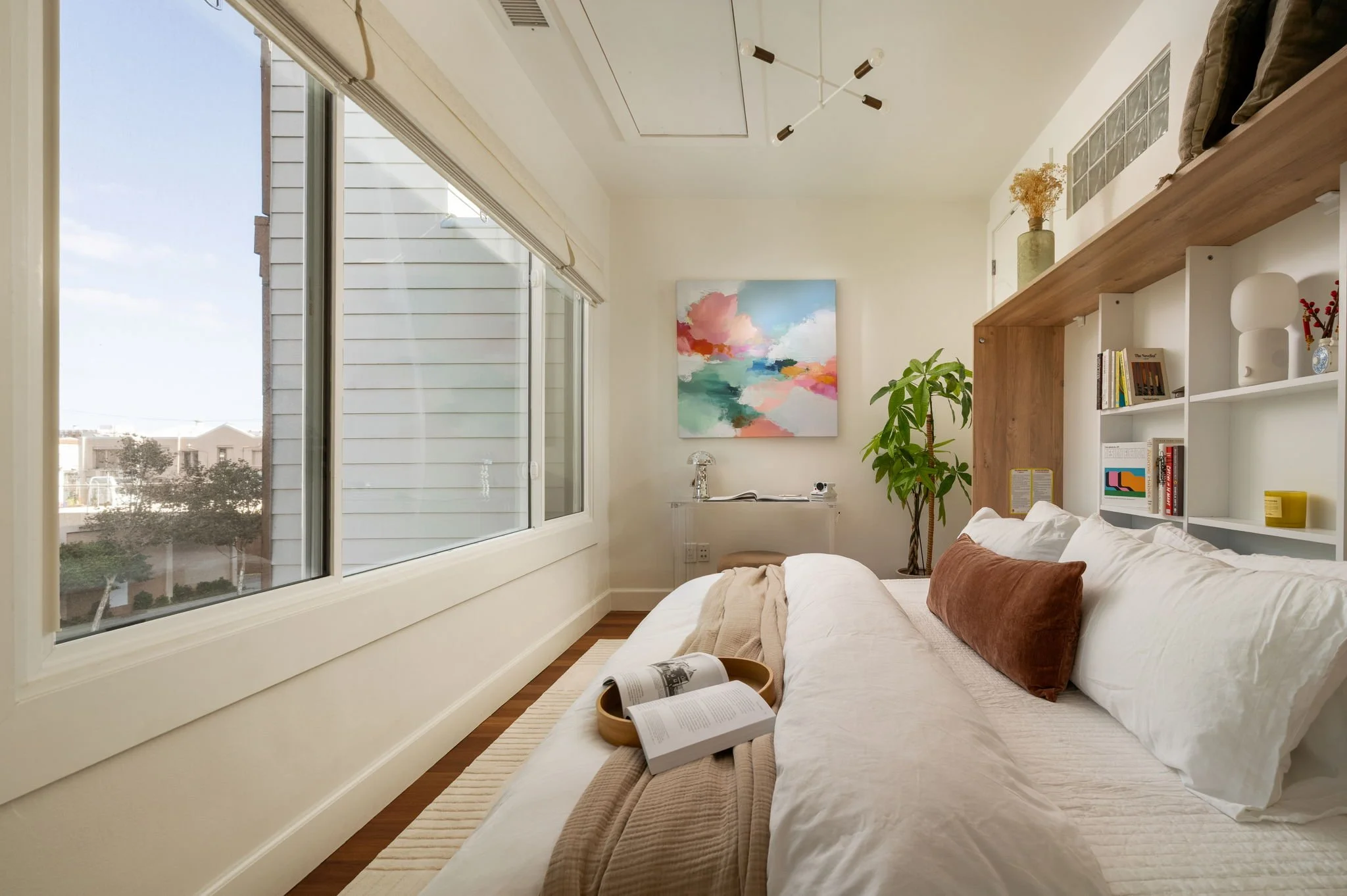 A bedroom with large windows showing a view of houses and trees outside. The bed has white bedding with beige and brown pillows. A tray with an open book and a magazine rests on the bed. There is a colorful abstract painting on the wall, a small tabl
