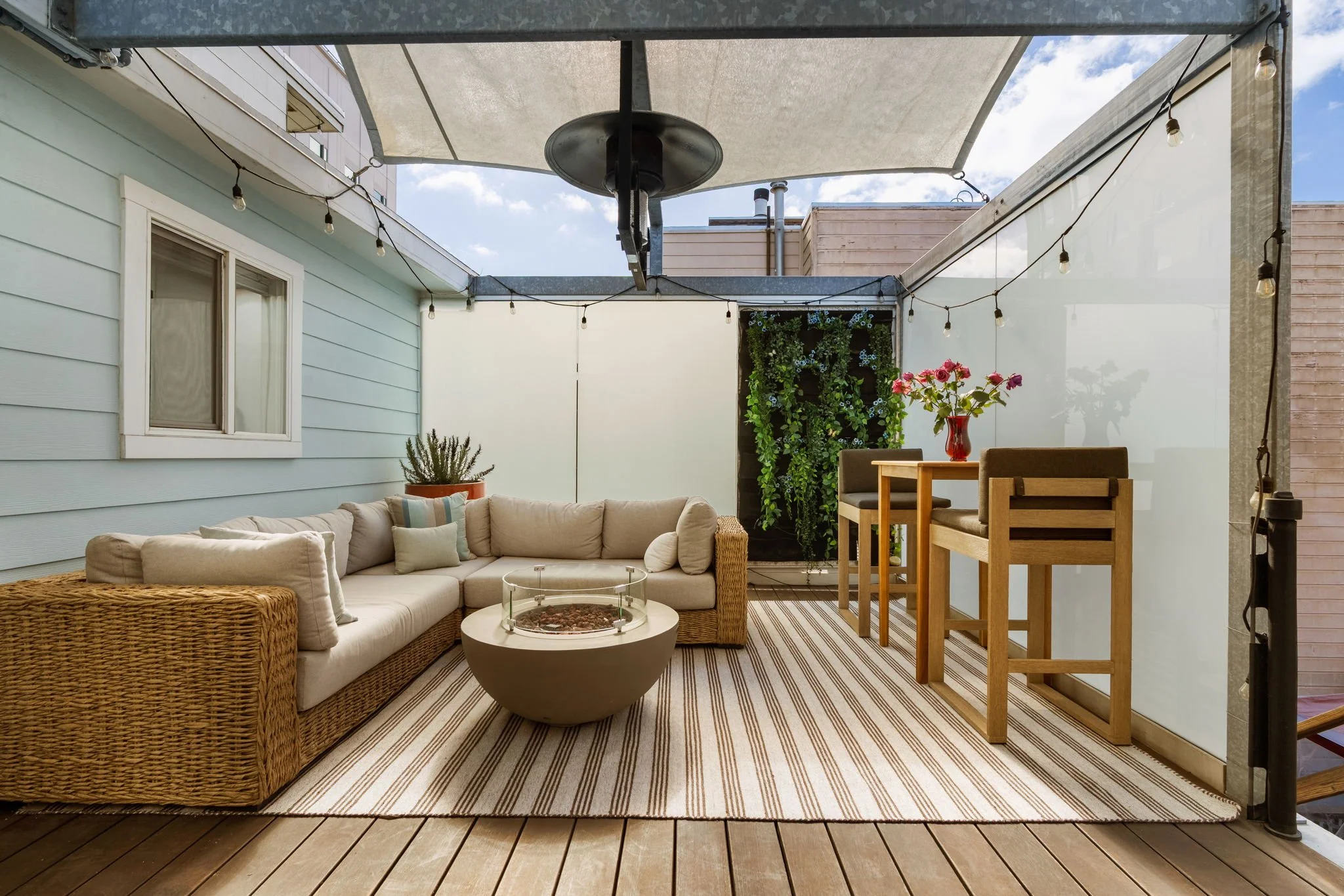 Outdoor patio with beige sectional sofa, wicker chair, glass fire pit, wooden high-top table and chairs, potted plant, vertical garden, string lights, striped outdoor rug, and clear sky.