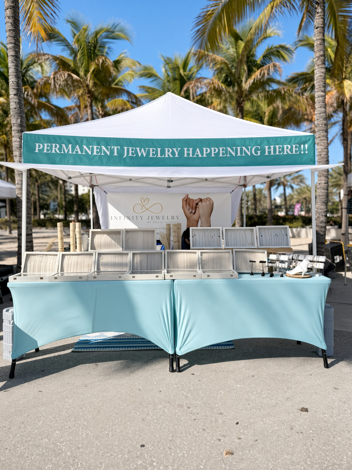 Infinity Jewelry permanent jewelry booth displaying chains and jewelry beneath a canopy surrounded by palm trees