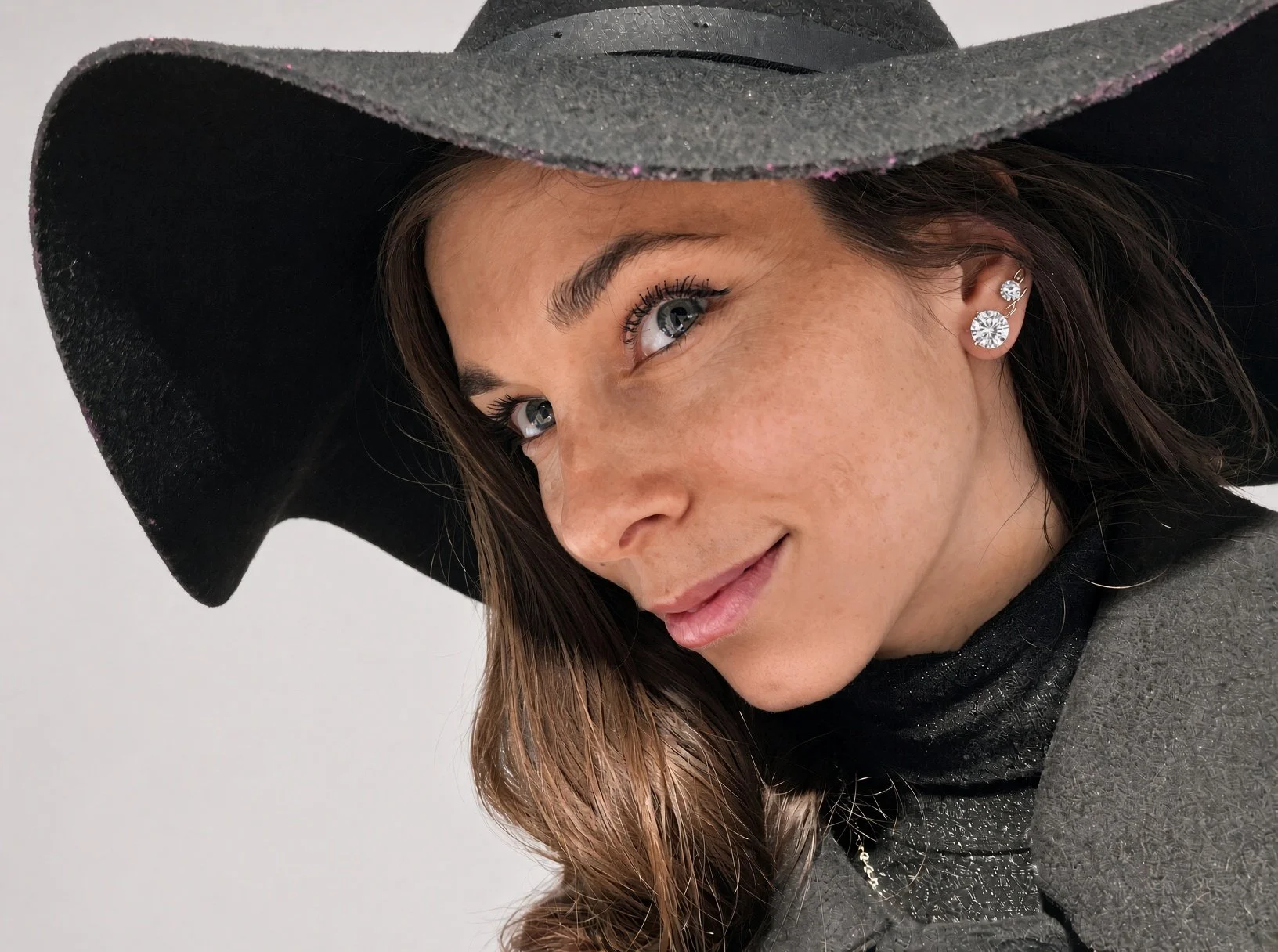 Close-up portrait of a woman wearing a black wide-brim hat, gray coat, black scarf, and sparkling ear jewelry.