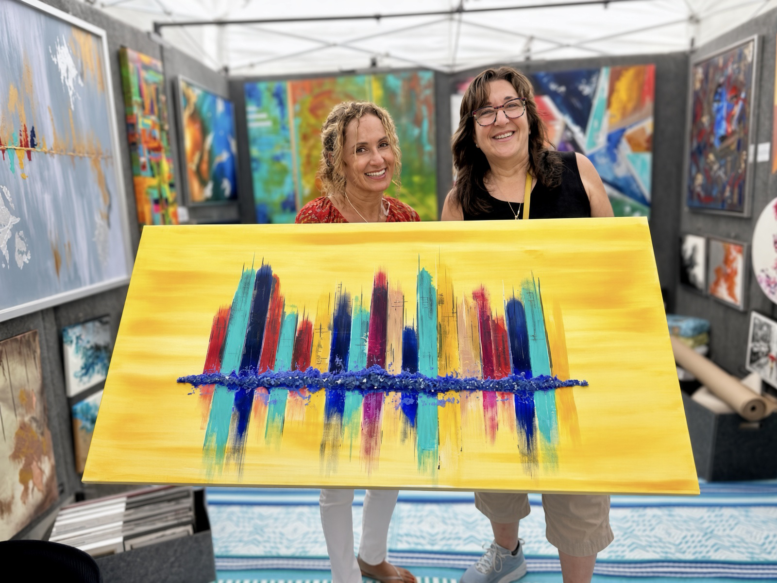 Two smiling female artists display a colorful abstract painting at an art exhibition booth.