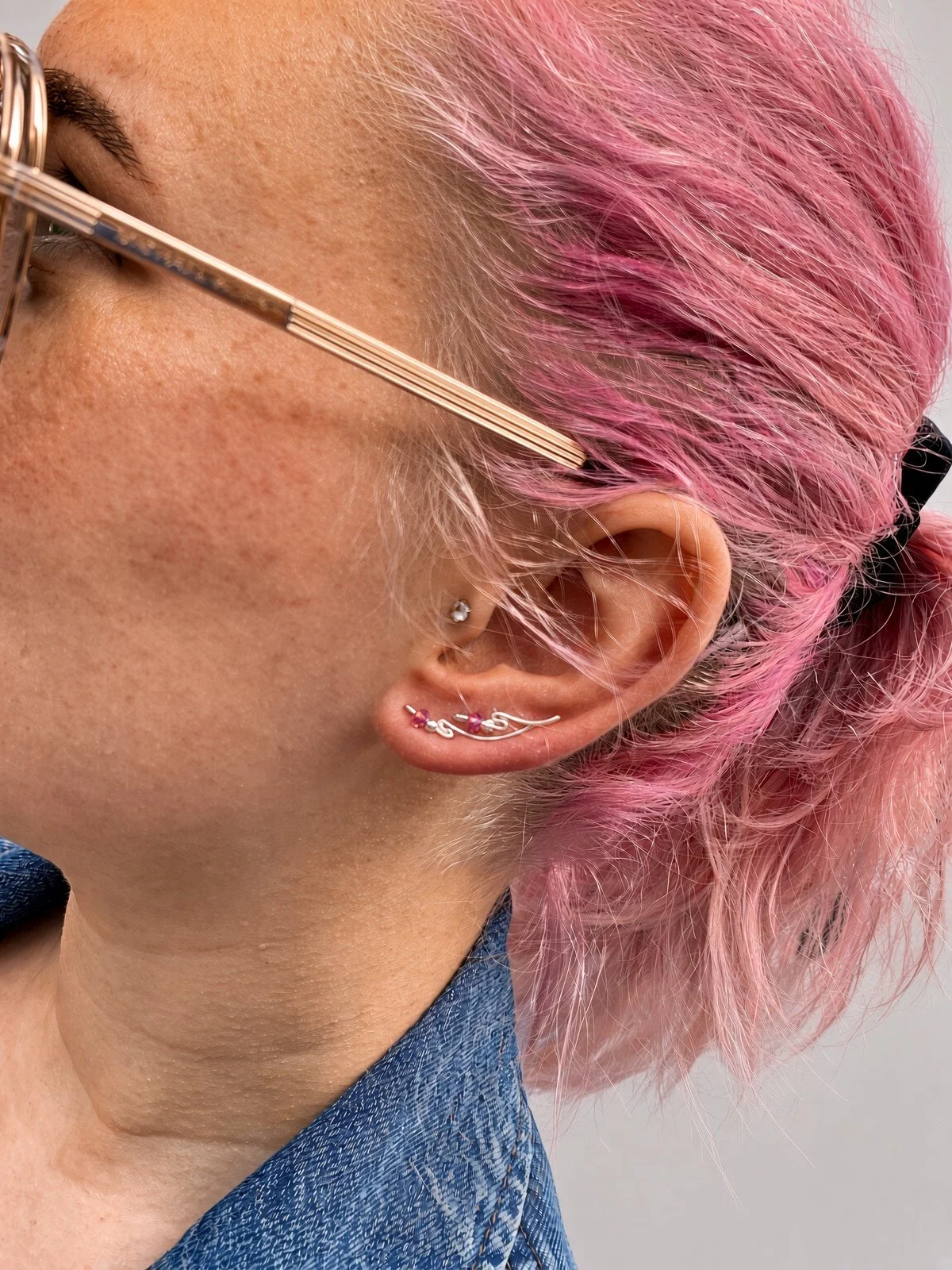 Close-up of a person with pink hair wearing gold eyeglasses and multiple silver ear piercings.