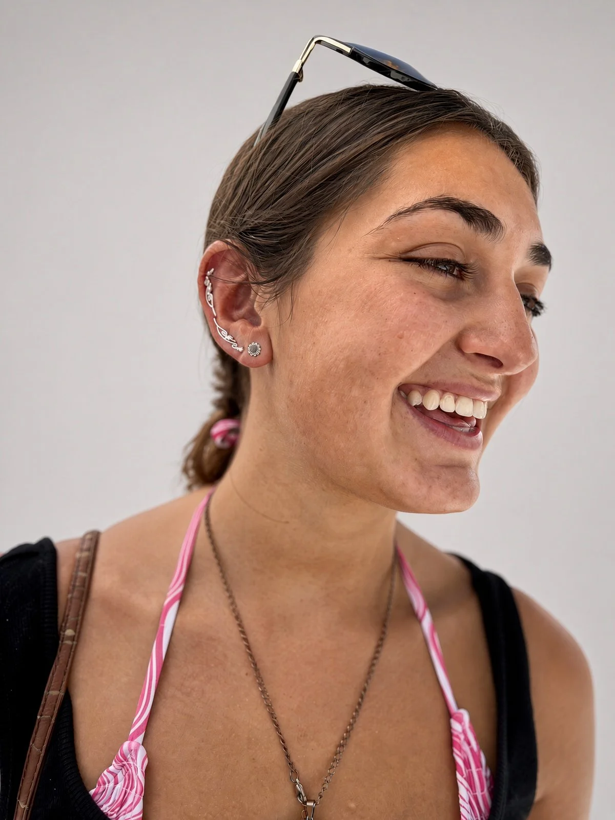 Smiling woman with sunglasses on her head and decorative ear jewelry, wearing a pink-and-white bikini top.