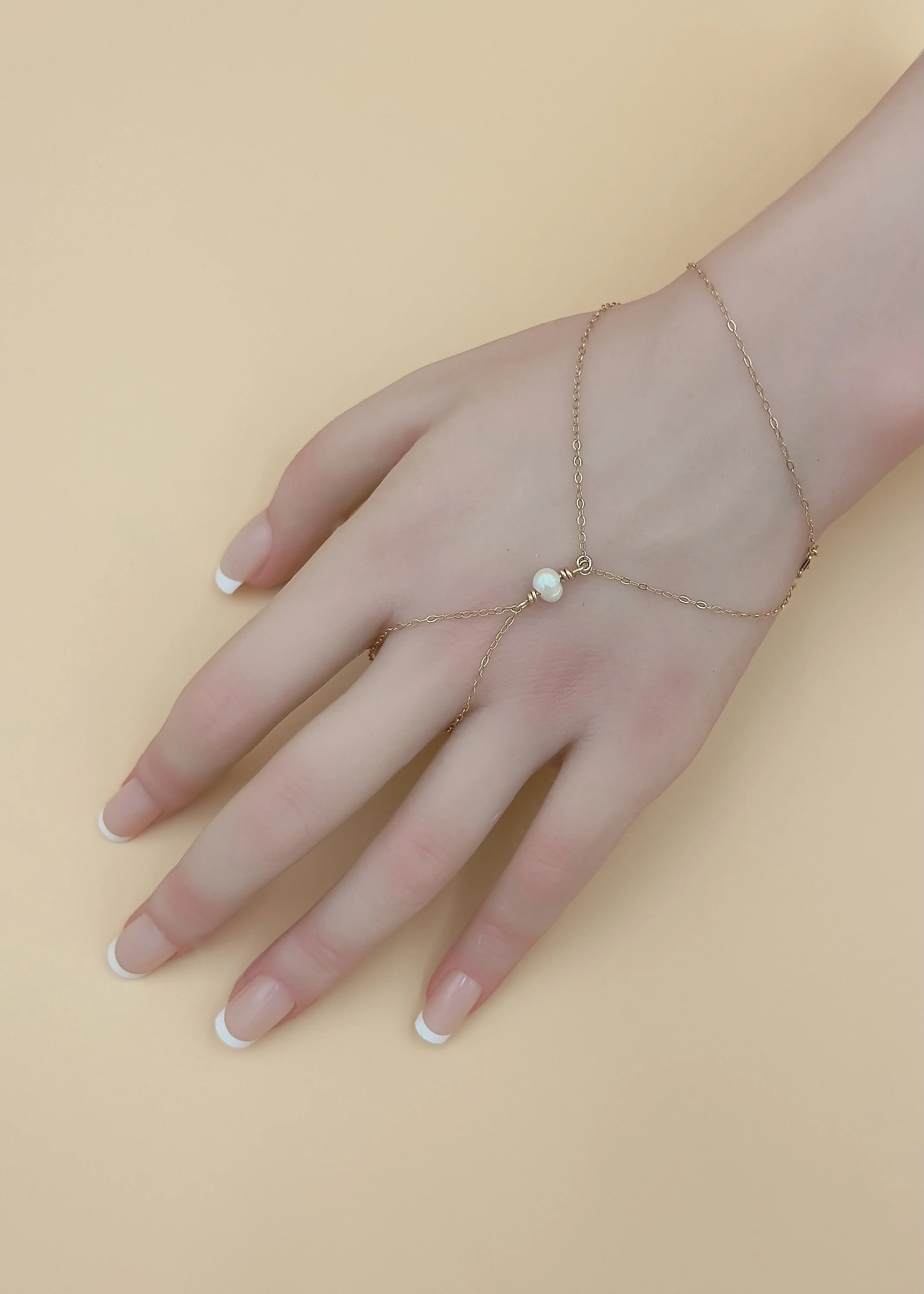 Delicate gold hand chain bracelet with a white pearl charm draped across a manicured hand.