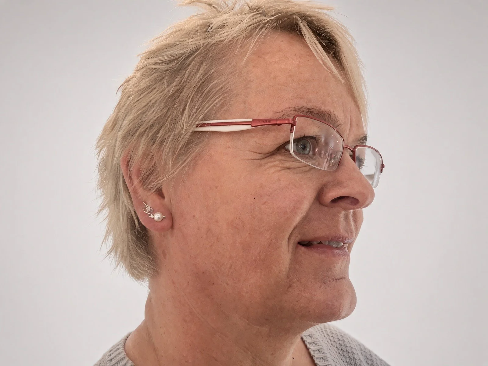 Close-up side profile of an older woman with short blonde hair, red-framed glasses, and pearl earrings