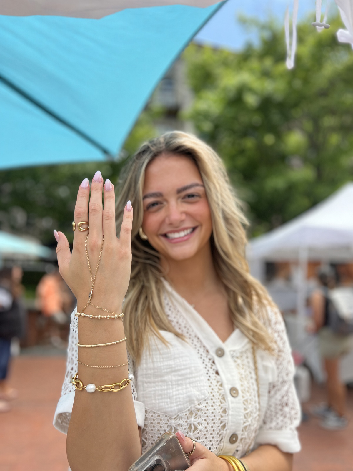Smiling woman showing her manicured hand and layered gold jewelry at an outdoor event.