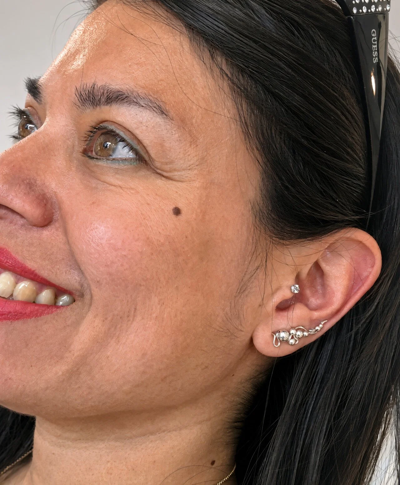 Close-up side profile of a smiling woman with dark hair, brown eyes, red lipstick, a cheek mole, and multiple ear piercings.