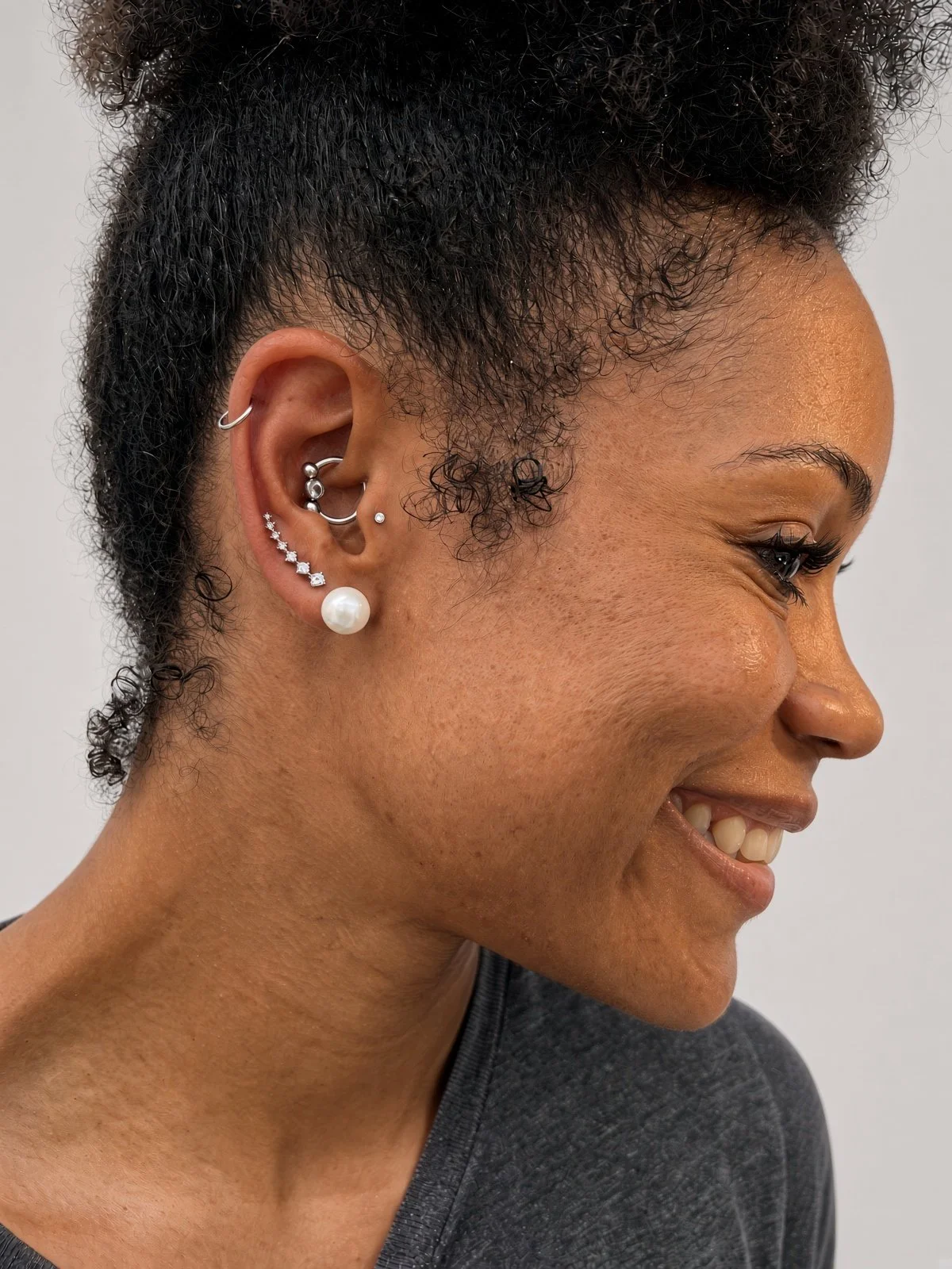 Close-up side profile of a Black woman with curly hair, dramatic eyelashes, and multiple silver ear piercings, including a large pearl earring.