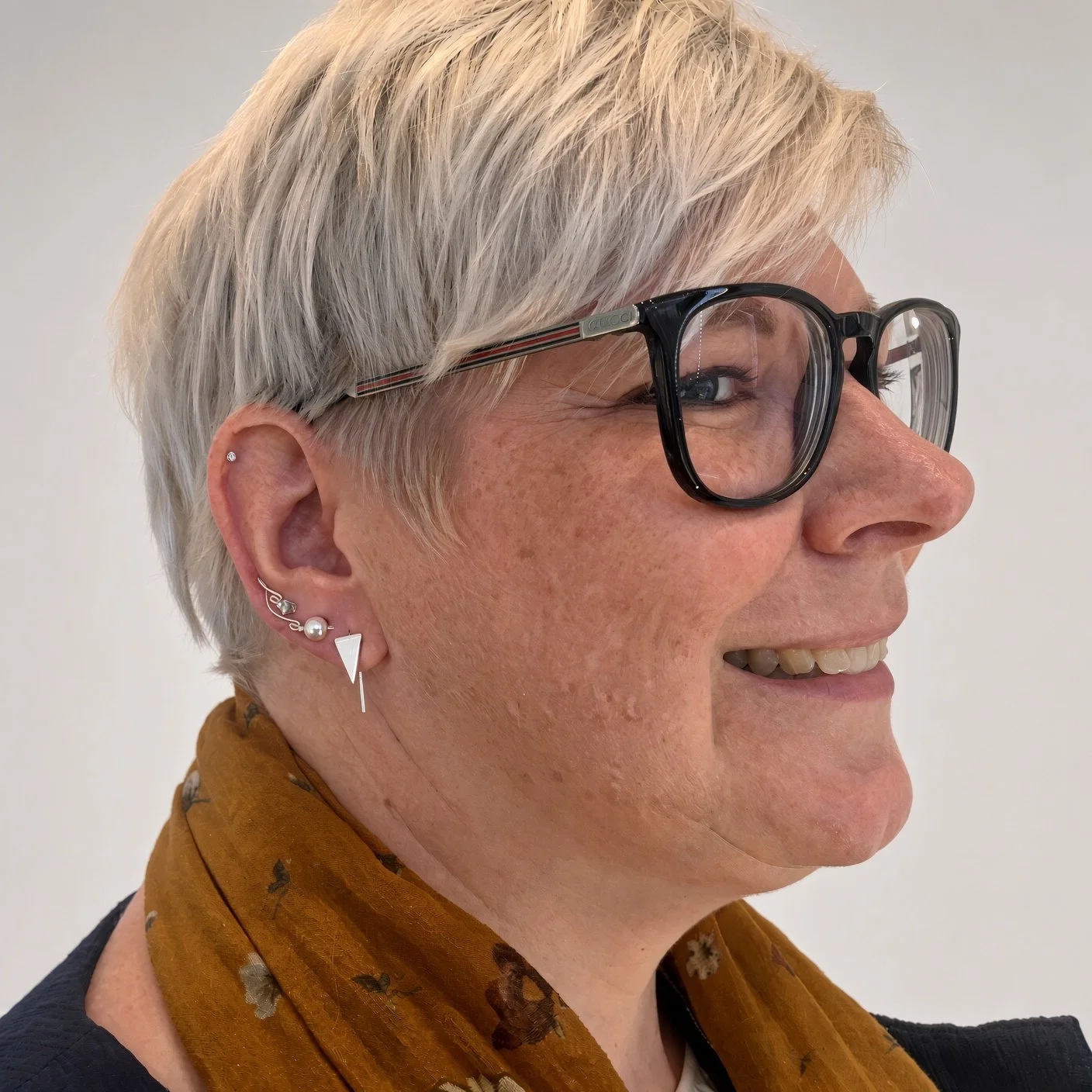 Side-profile portrait of an older woman with short silver hair, black eyeglasses, layered earrings, and a mustard floral scarf