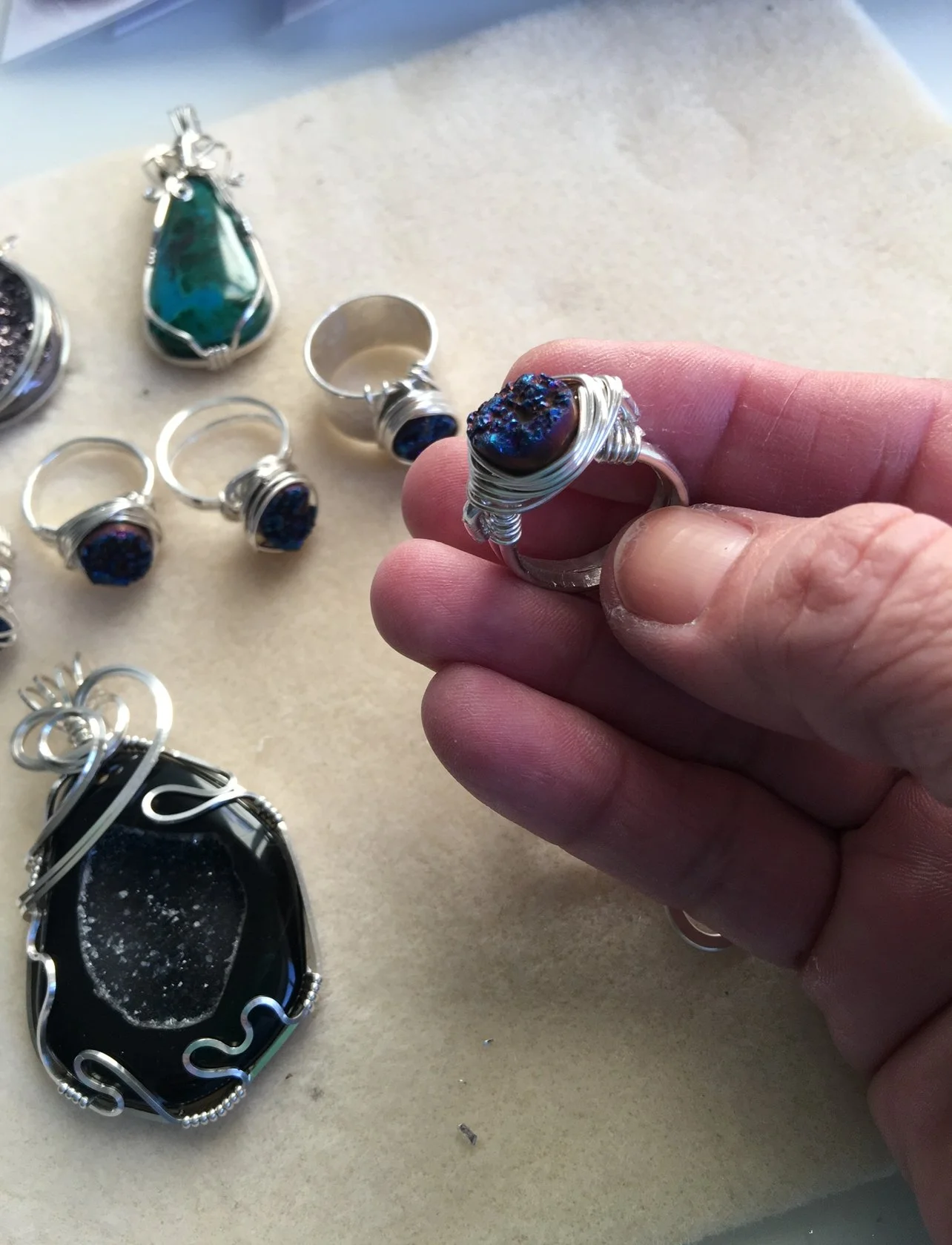Hand holding a handmade silver wire-wrapped ring with a dark blue textured stone, surrounded by gemstone jewelry.