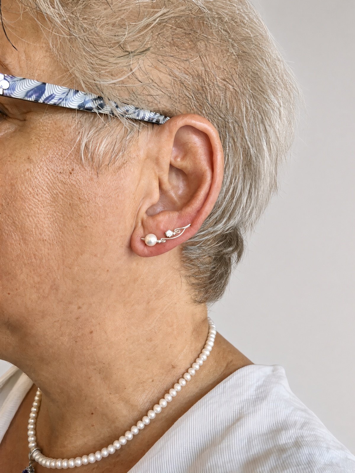 Close-up of an older woman wearing patterned glasses, pearl earrings, and a pearl necklace.