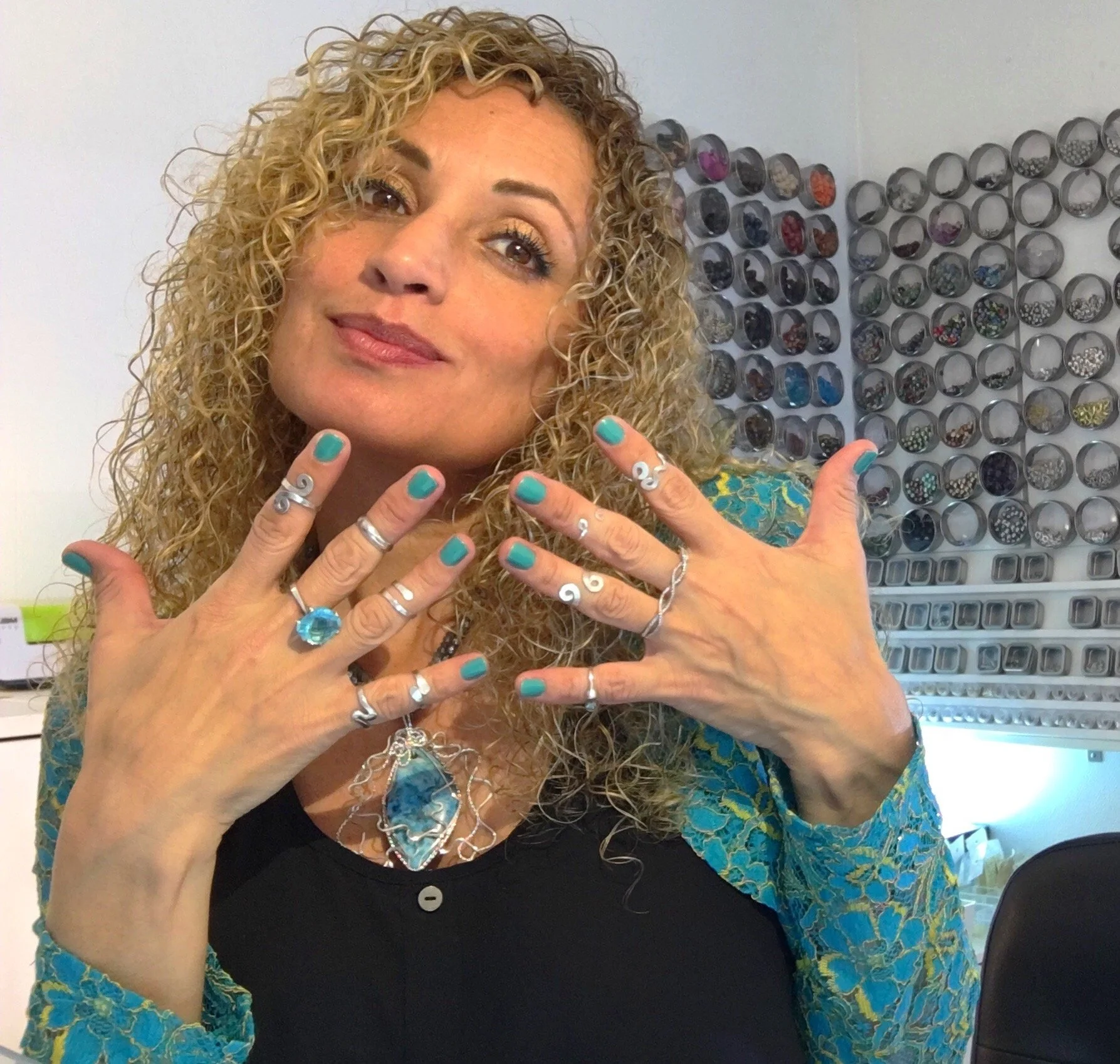 Woman with curly blonde hair, turquoise nail polish, multiple rings, and a large blue pendant necklace posing indoors