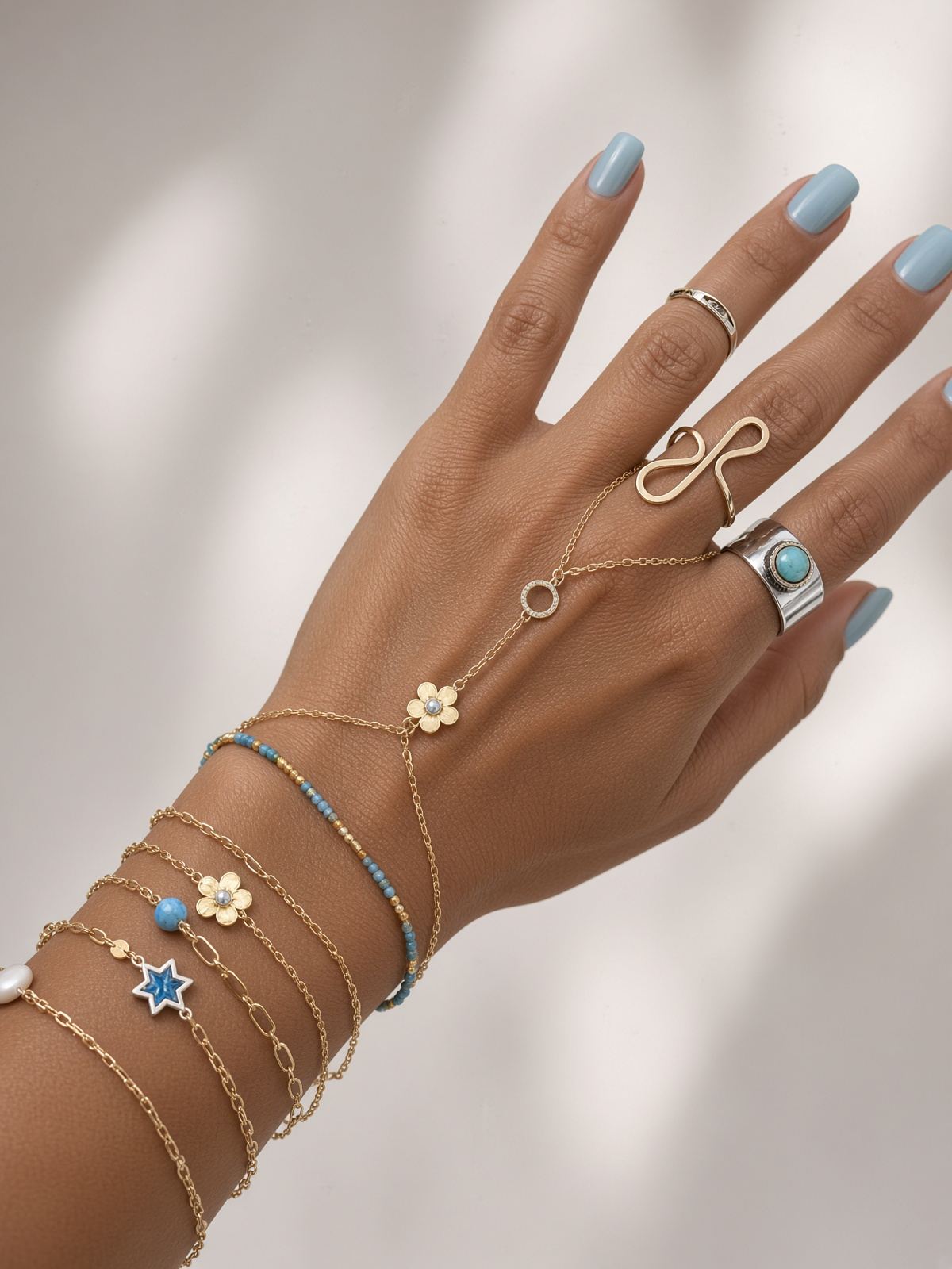 Close-up of a hand with light blue manicured nails wearing layered gold bracelets, rings, and a delicate hand chain with turquoise and floral charms.