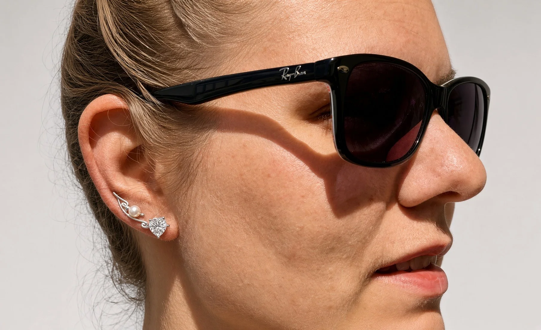 Close-up side profile of a woman wearing black Ray-Ban sunglasses and decorative pearl and heart-shaped earrings