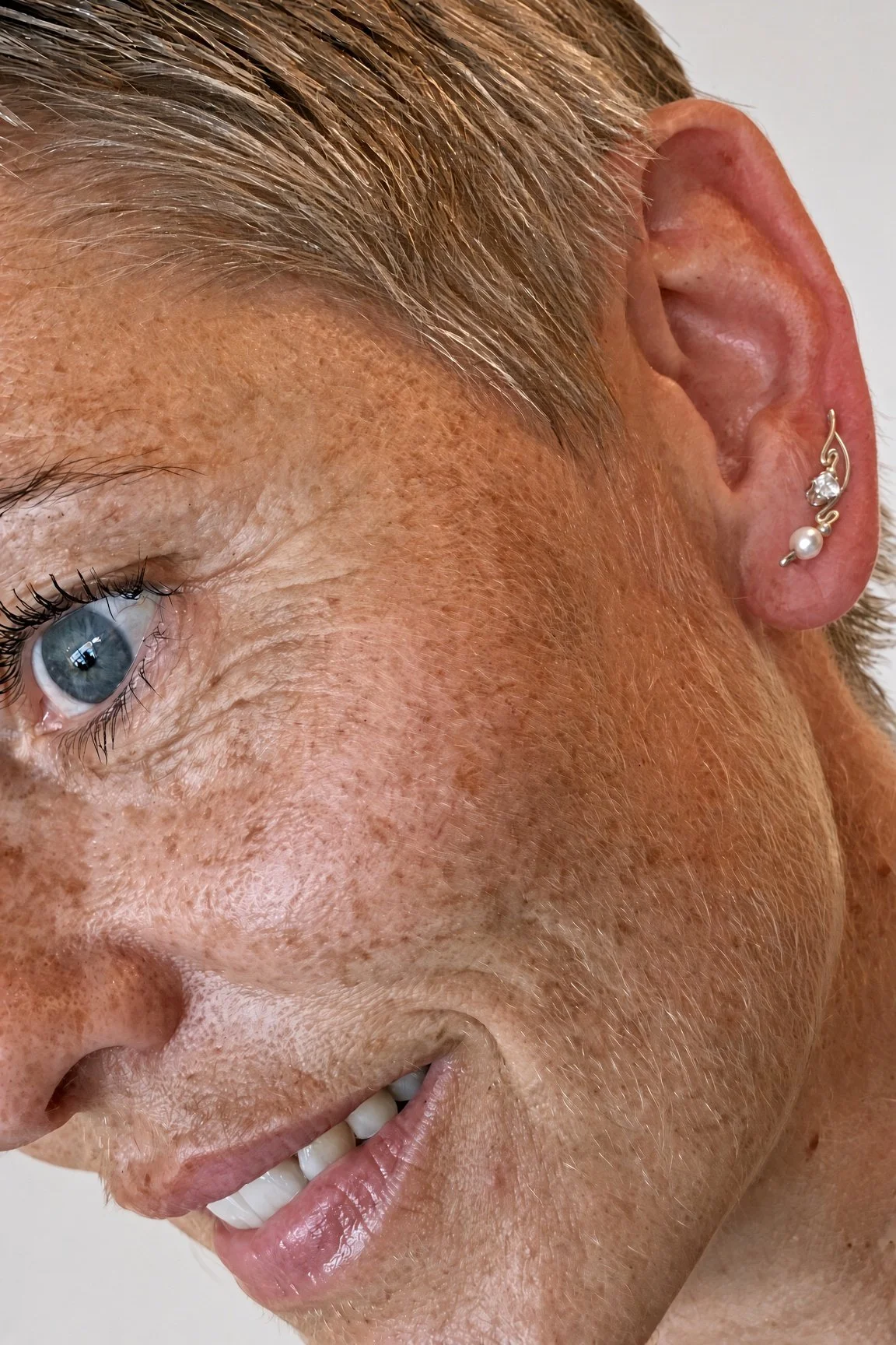Close-up side profile of an older woman with blue eyes, gray hair, freckles, and pearl-and-crystal earrings