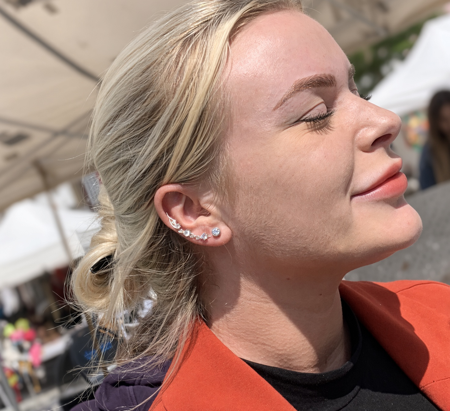 Close-up side profile of a blonde woman wearing multiple silver ear piercings and an orange jacket outdoors