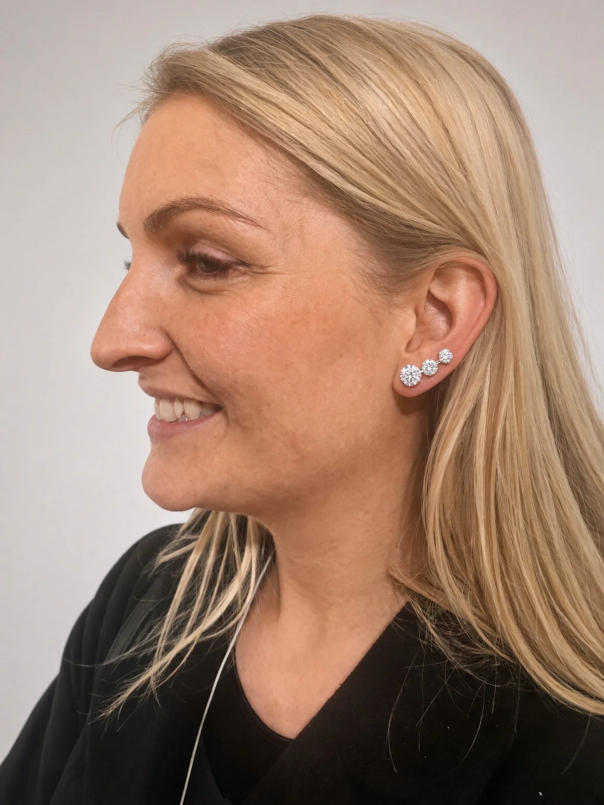 Side-profile portrait of a smiling blonde woman wearing three diamond stud earrings