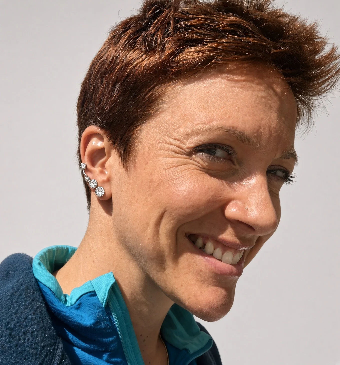 Close-up portrait of a smiling person with short brown hair, multiple silver ear piercings, and a blue jacket
