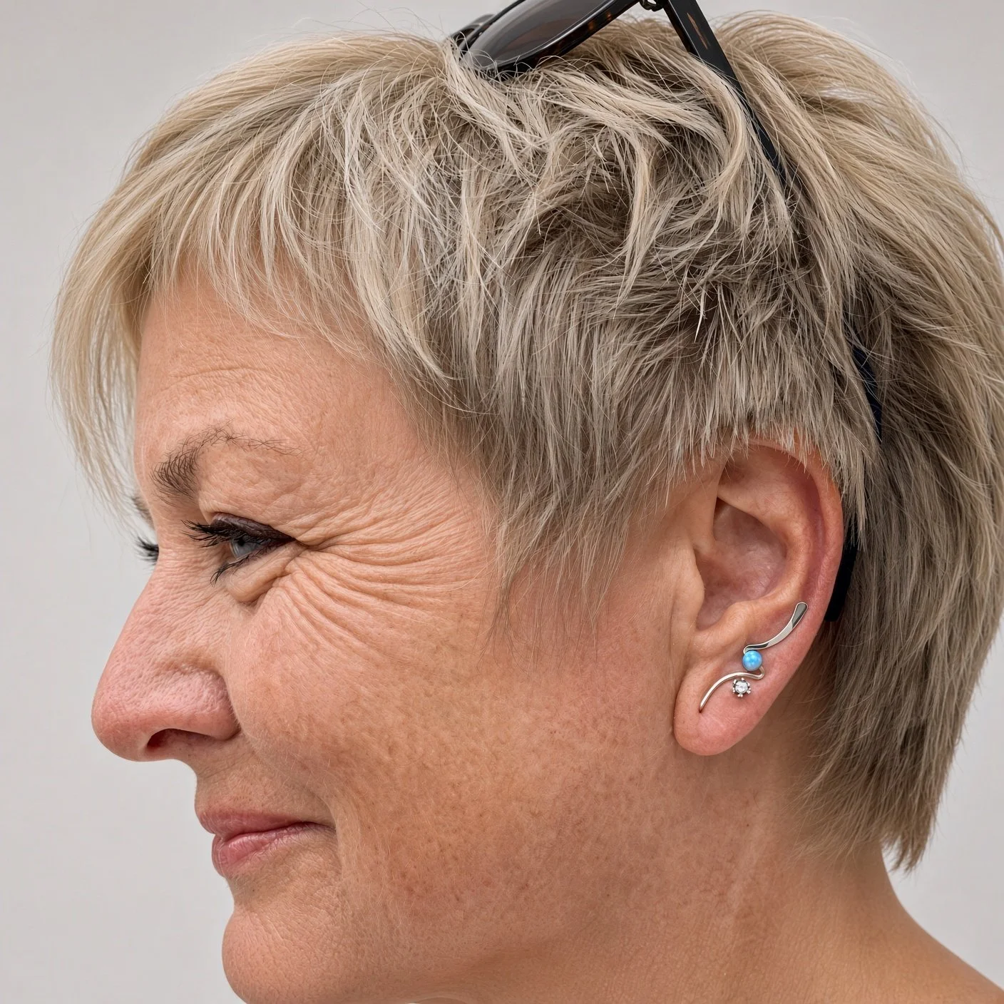 Close-up side profile of an older woman with gray hair, glasses, earrings, and visible facial wrinkles