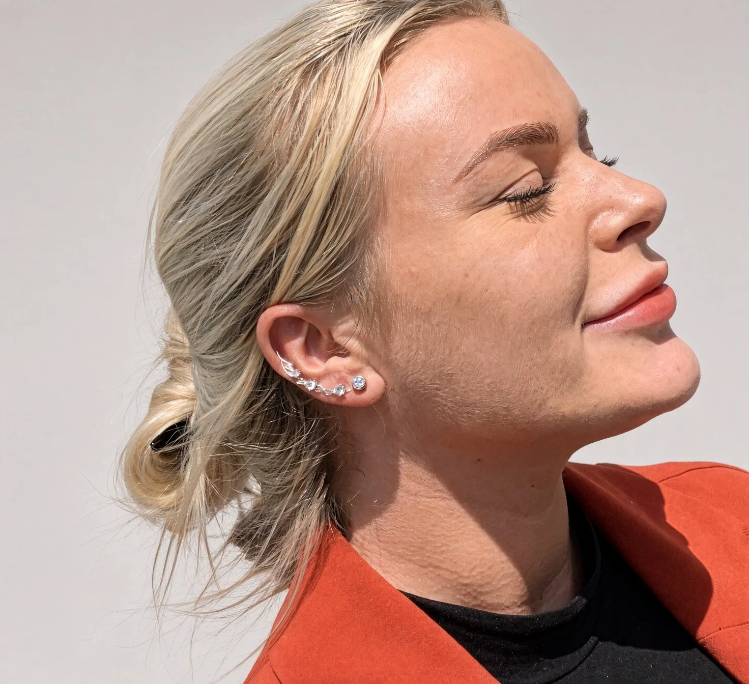 Close-up side profile of a blonde woman wearing multiple silver ear piercings and an orange jacket