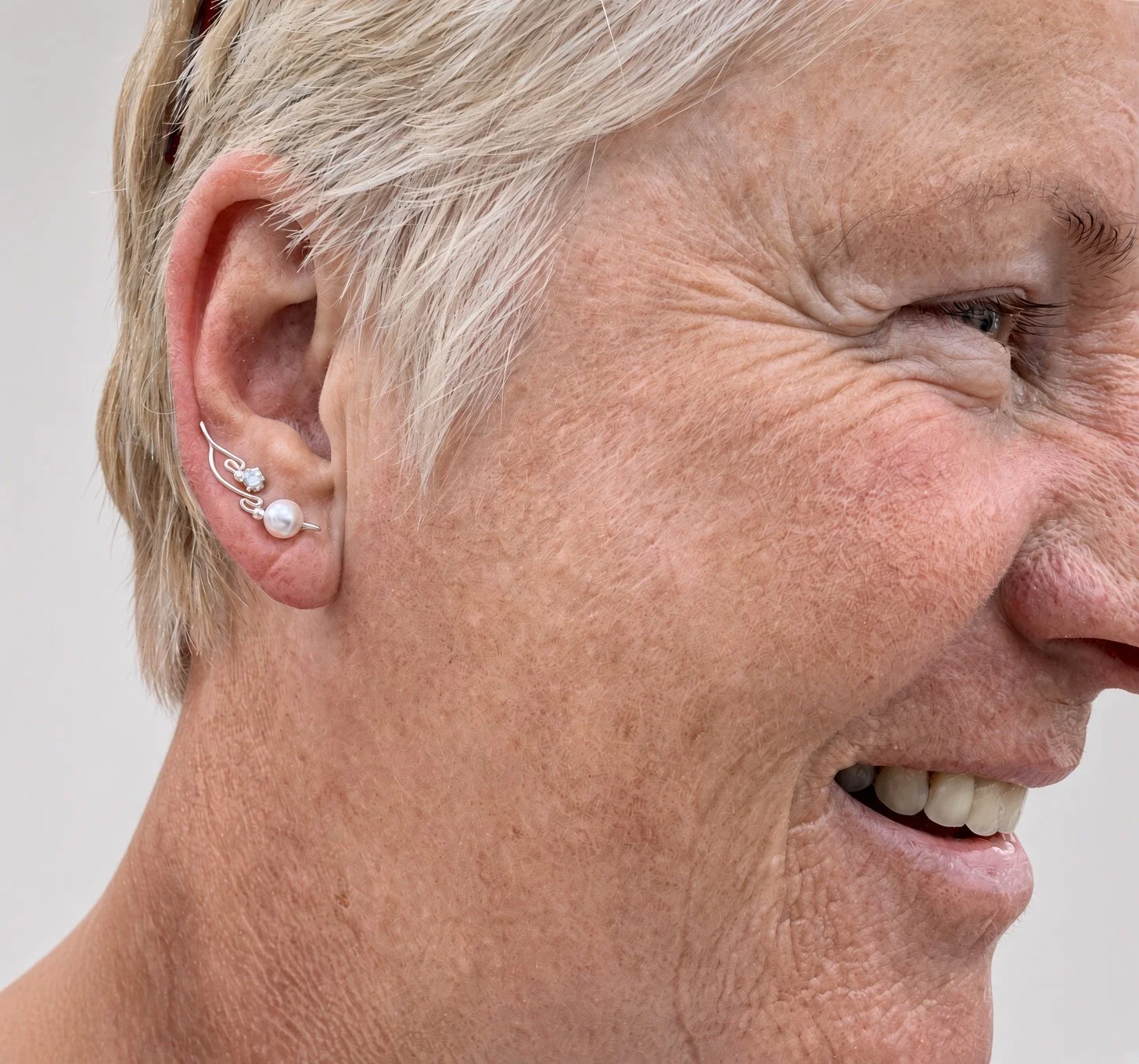 Close-up profile of an elderly woman wearing pearl and silver earrings, smiling and squinting.