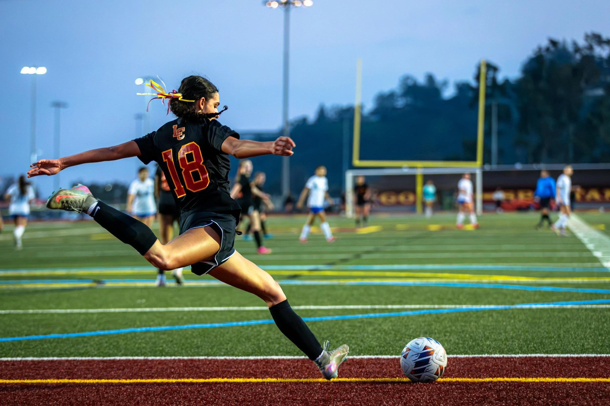A female soccer player wearing a black jersey with the number 18 is kicking a soccer ball on the field during dusk or early evening, with other players in the background.