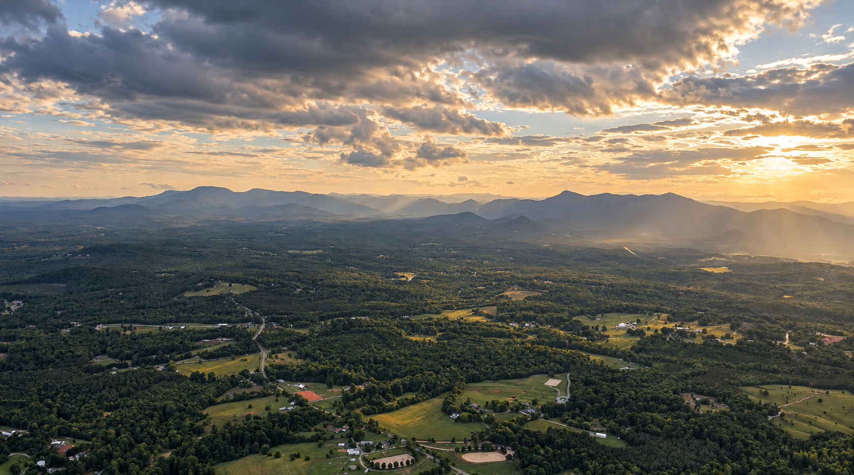 Aerial view of rolling green farmland and forests beneath a dramatic sunset sky, with layered mountain ranges in the distance