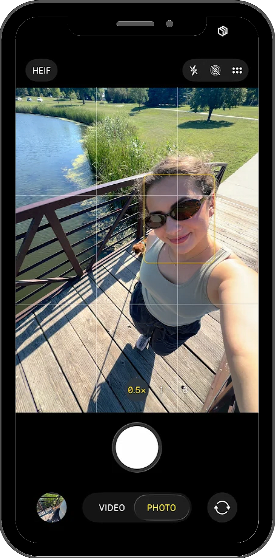 Smartphone camera screen showing a woman in sunglasses taking a selfie on a wooden footbridge beside a pond.