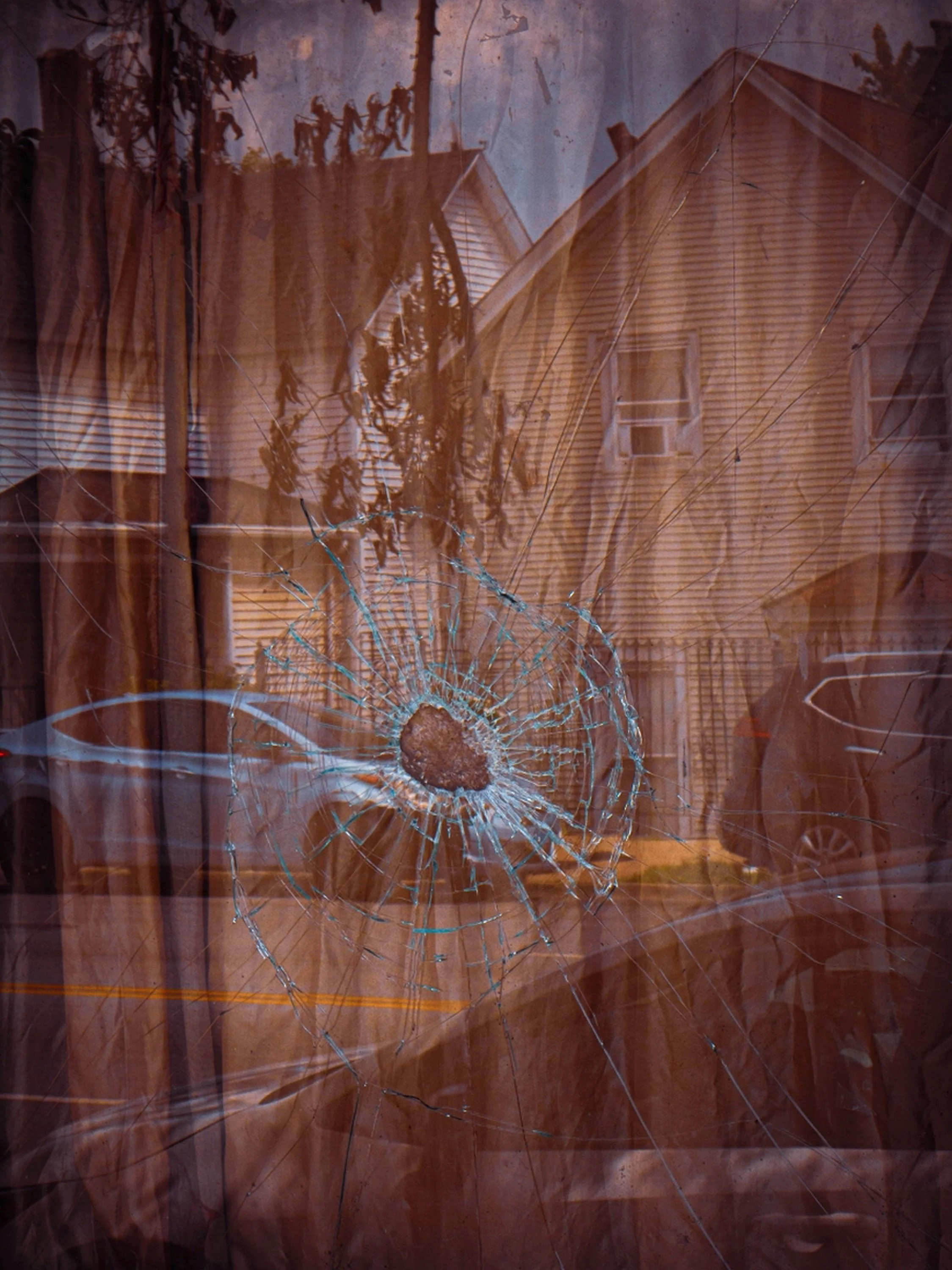 Broken glass window with a central hole and radiating cracks, reflecting a wooden house exterior with windows and trees outside.