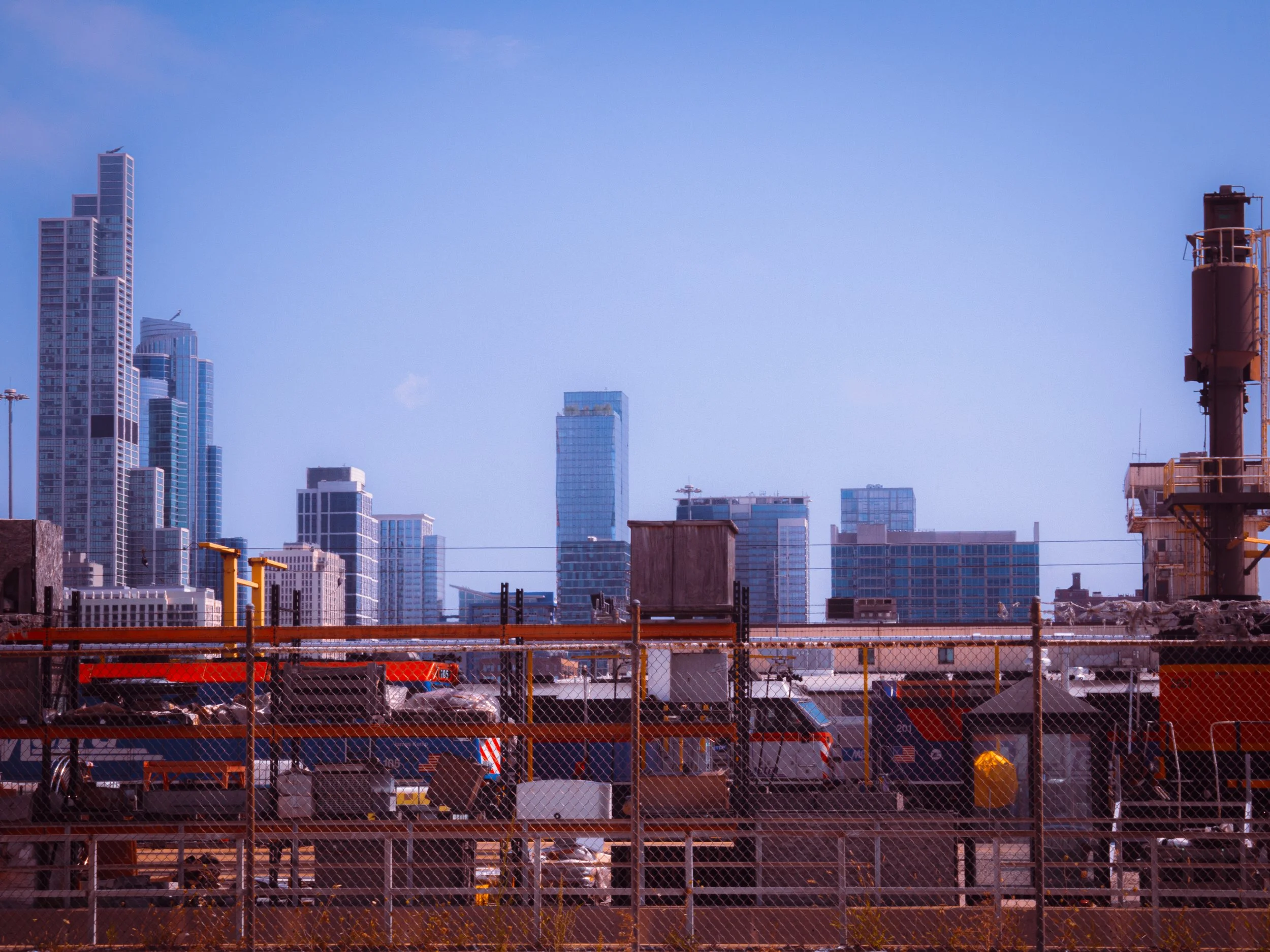 View of a city skyline with tall modern buildings under a clear blue sky. In the foreground, there is a construction site with orange safety barriers, fencing, and construction equipment.