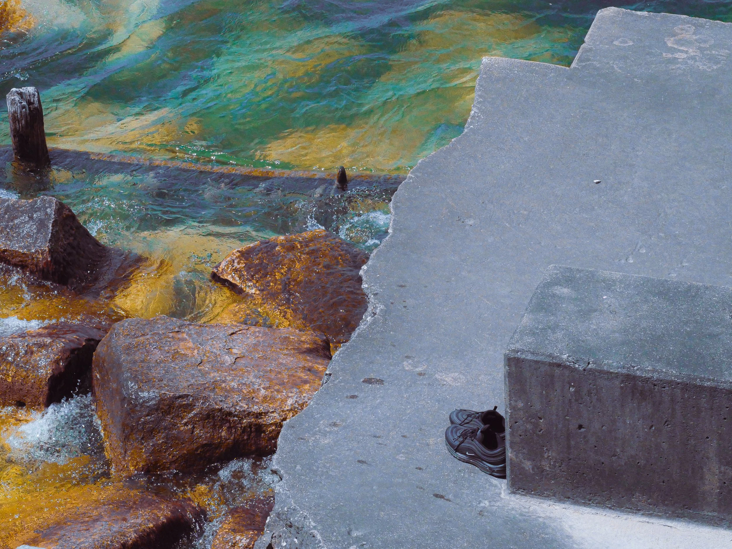 Photo taken from a person standing on concrete stairs by the water. The person's black sneakers with gray soles and black pants are visible at the bottom of the photo. The water is clear with visible stones and rocks underneath, and some wooden posts