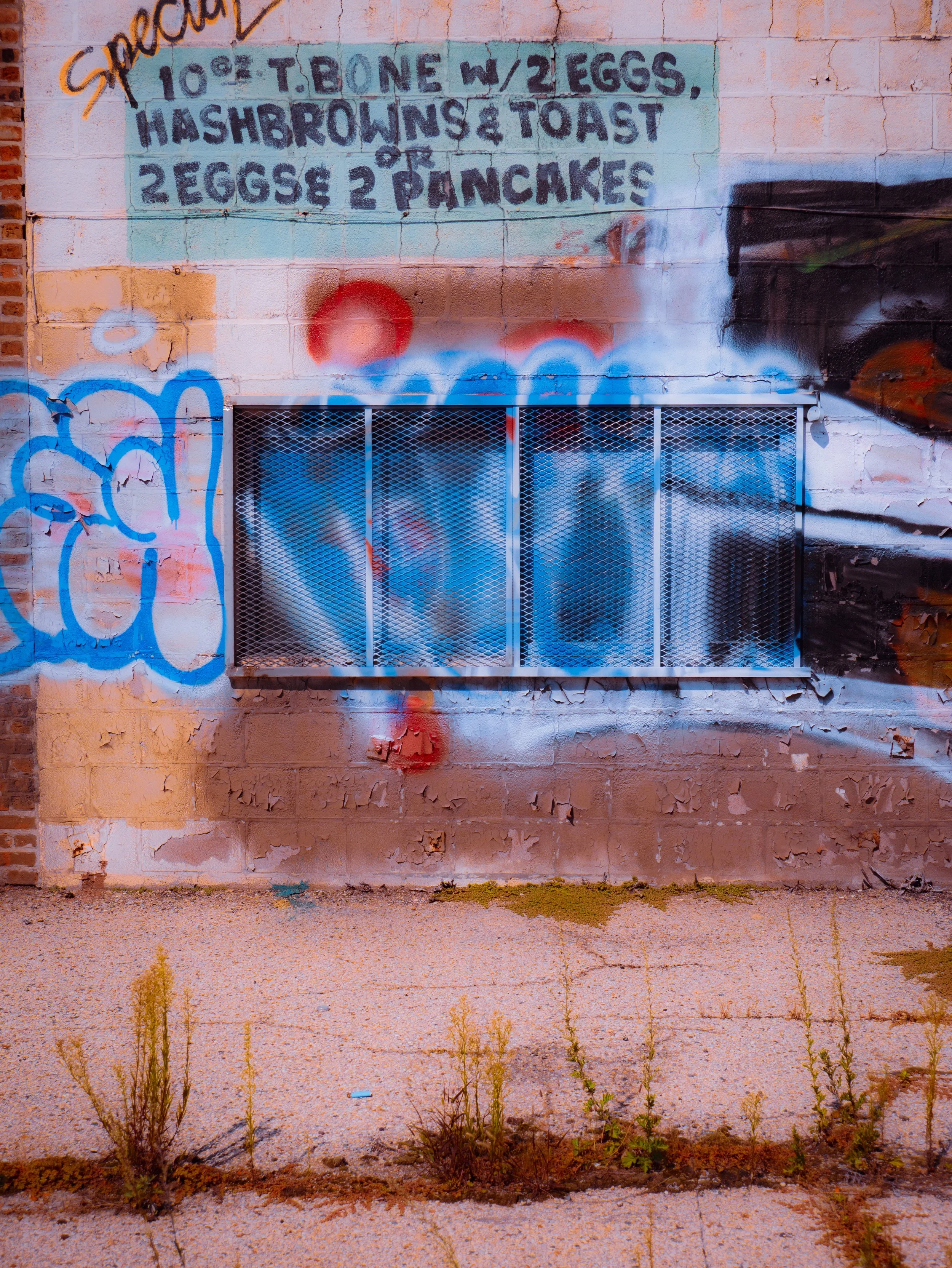 A graffiti mural on a brick wall featuring a menu with prices and items like pancakes and eggs, painted in a street art style. The wall shows peeling paint and some graffiti art, with weeds growing at the base of the wall.