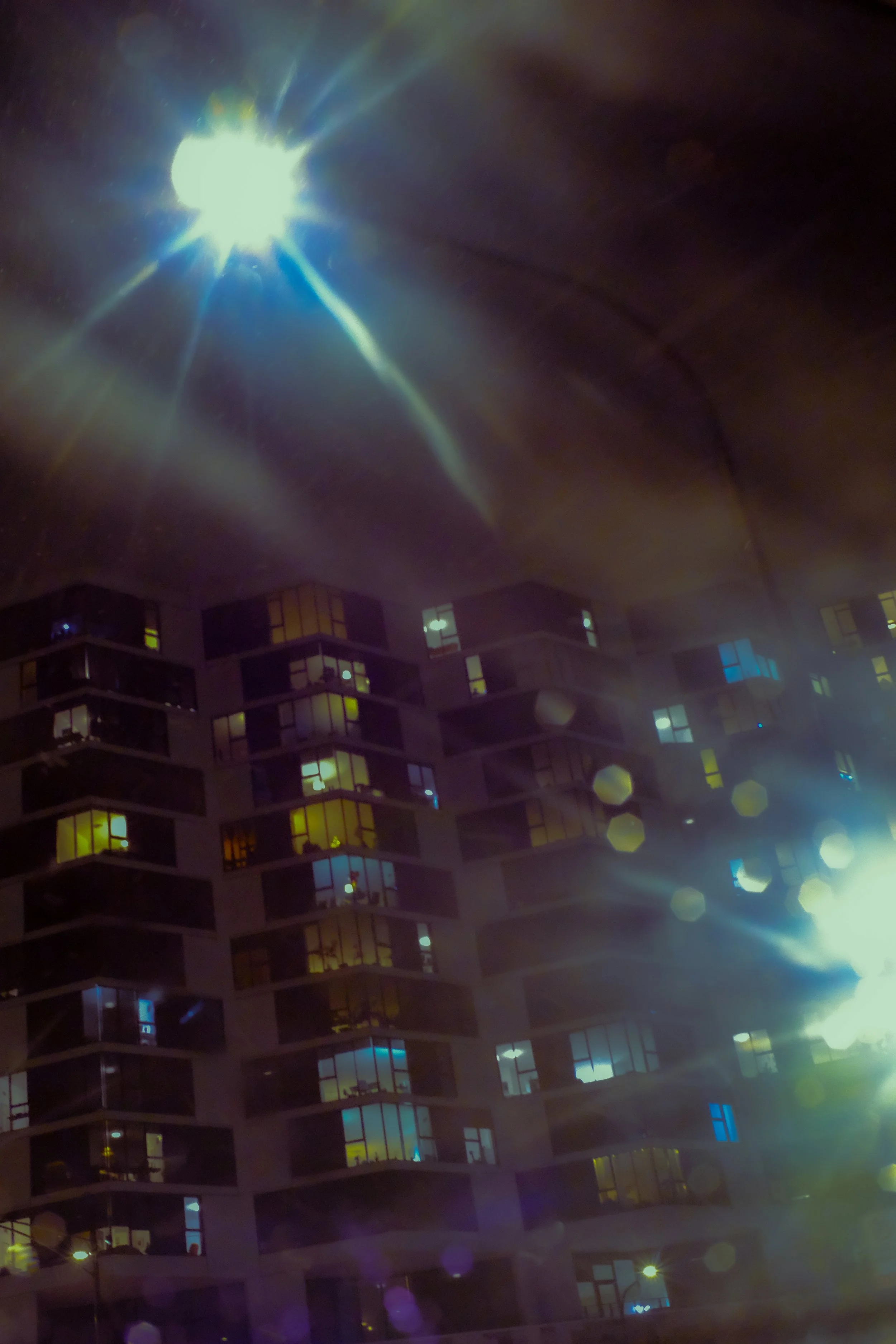Nighttime city scene with a tall modern building illuminated by interior lights and a bright light source in the sky creating starburst effects.