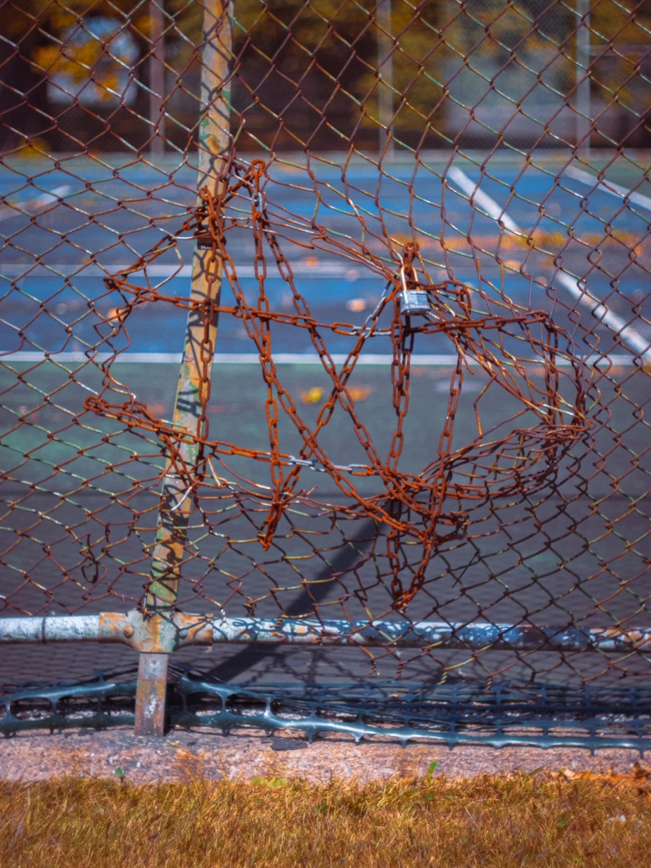 A rusted chain-link fence with a padlock, preventing access to an outdoor tennis court amidst autumn foliage.