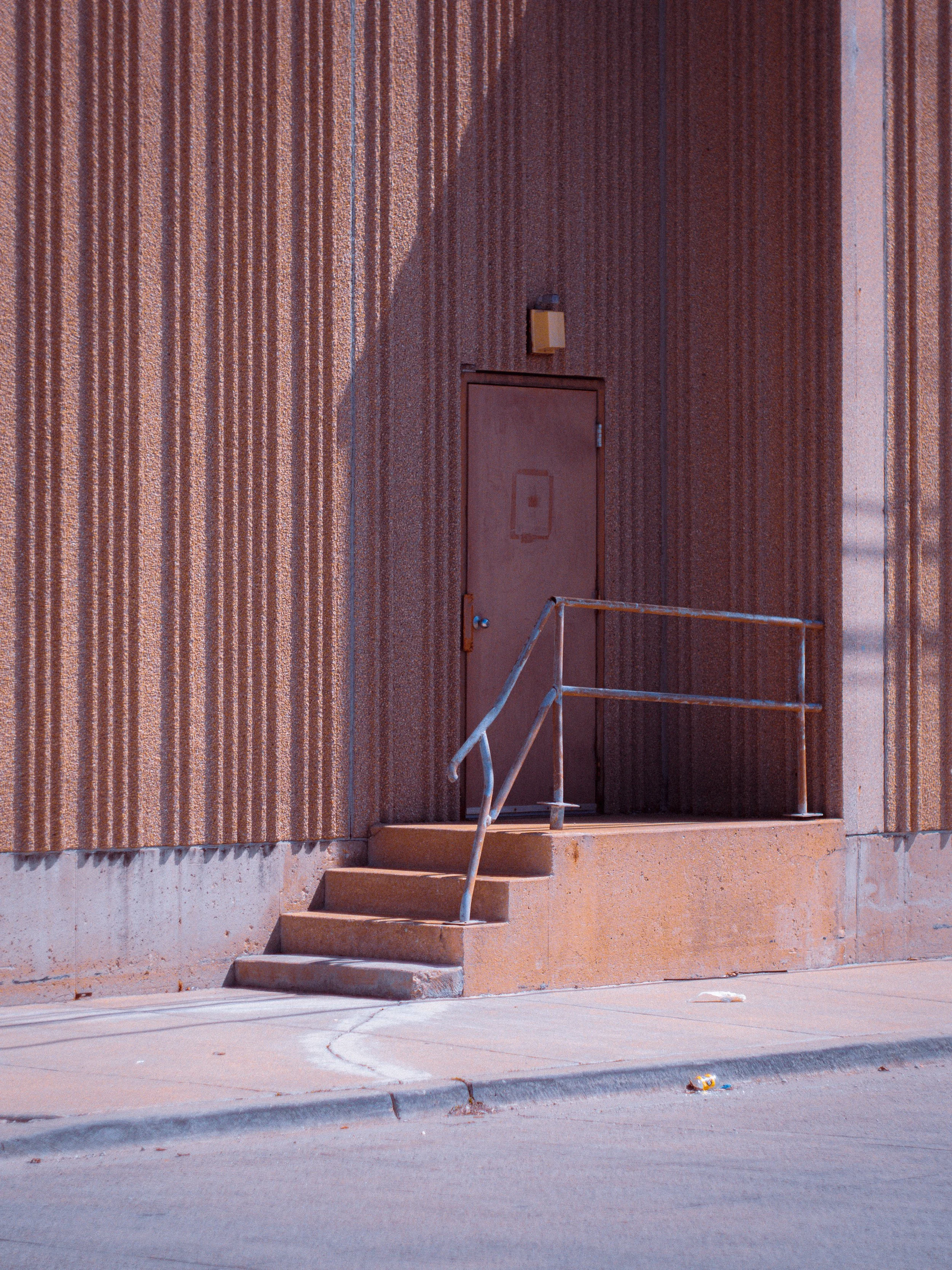A brown door with a small window, outdoor steps with metal railing, and a textured wall with vertical stripes. There is a small trash item near the curb.