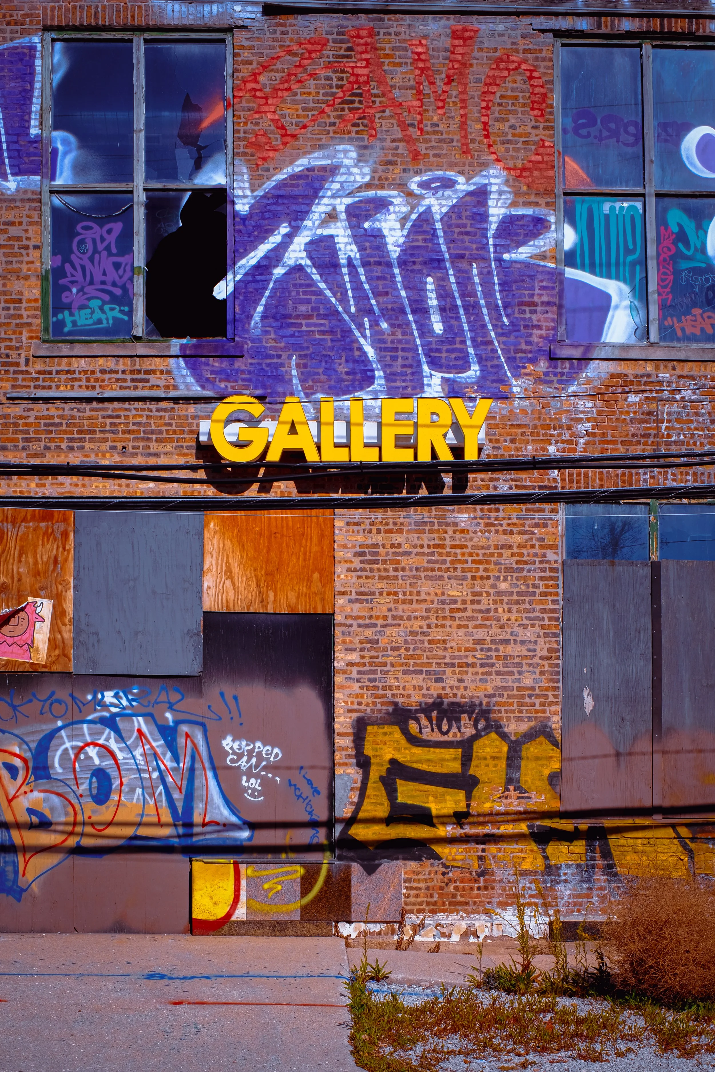 The image shows a brick wall with graffiti and a large illuminated yellow sign that reads 'GALLERY'. There are two broken and graffitied window shutters or panels, and some small plants are growing in front of the panels on the ground. The wall also 