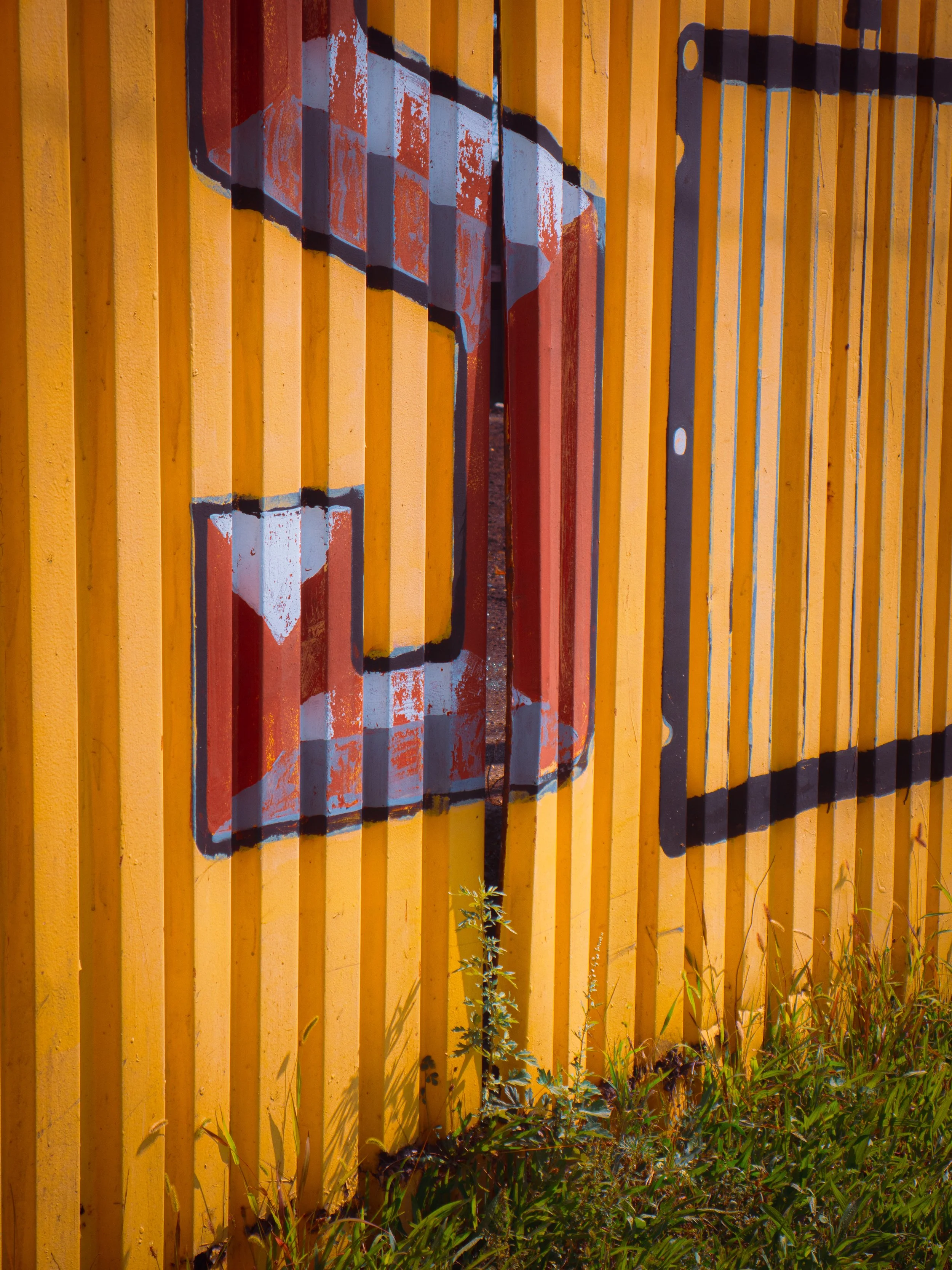 Yellow wooden fence with a colorful painted design, plants and grass growing at the base.