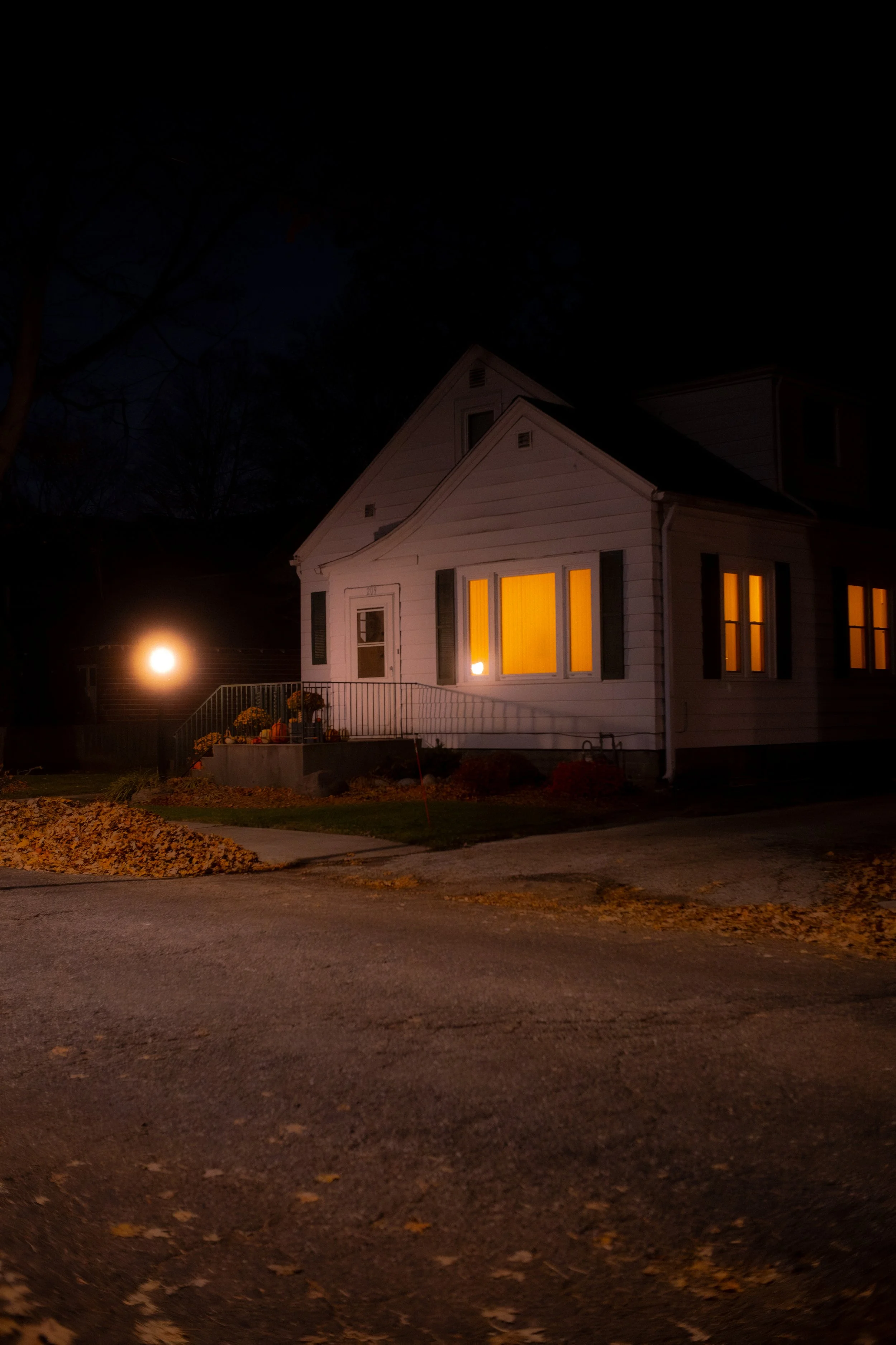 A house at night with warm yellow lights shining from its windows, a small porch with pumpkins and flowers, a driveway, and a tree with fallen leaves in the foreground. A bright streetlight is visible in the background.