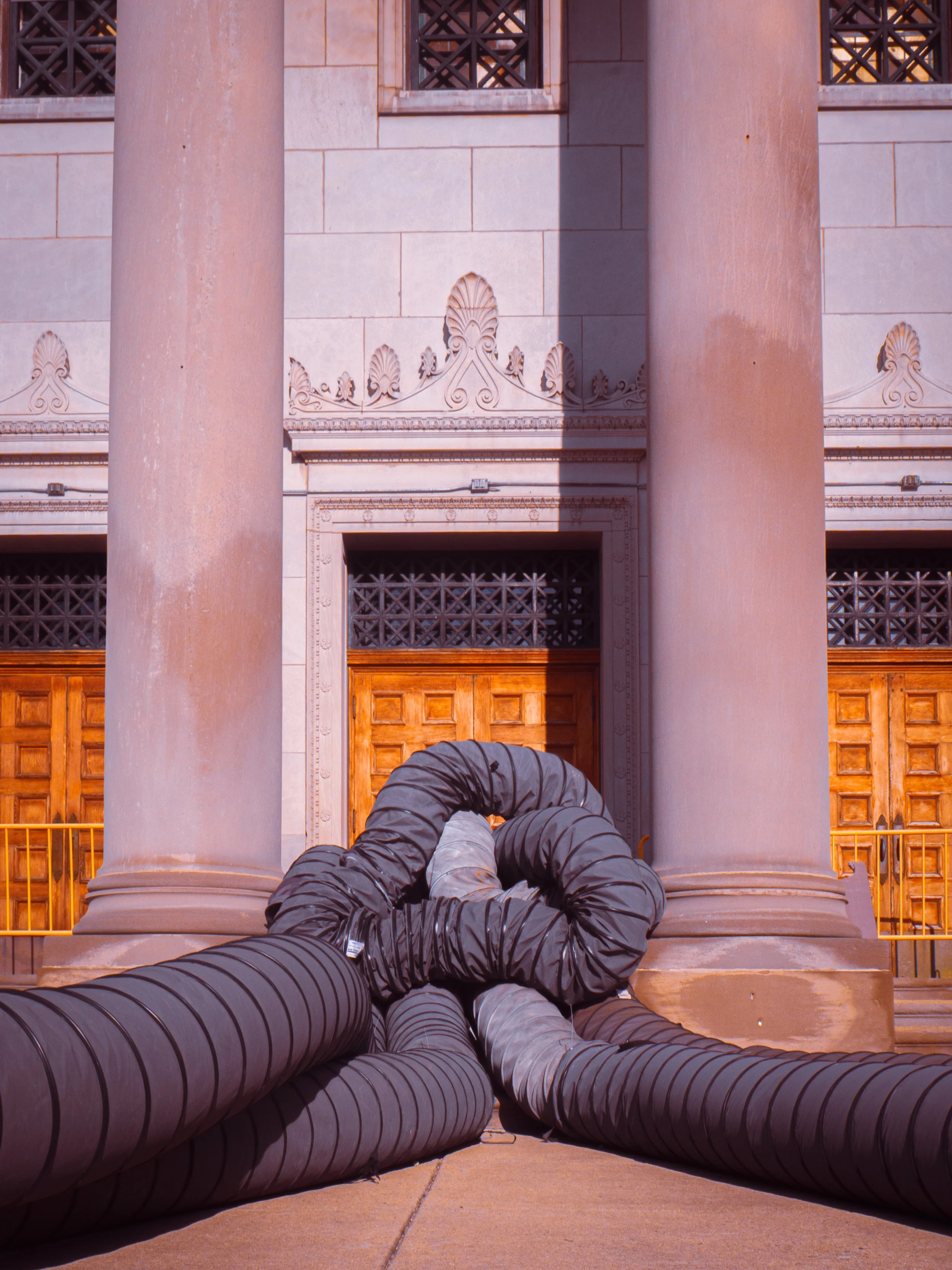 A person with dark clothing and a jacket is lying on the ground at the base of a building with large pink columns and ornate architectural details. They are positioned with their head towards the building and their legs extended outward, with multipl
