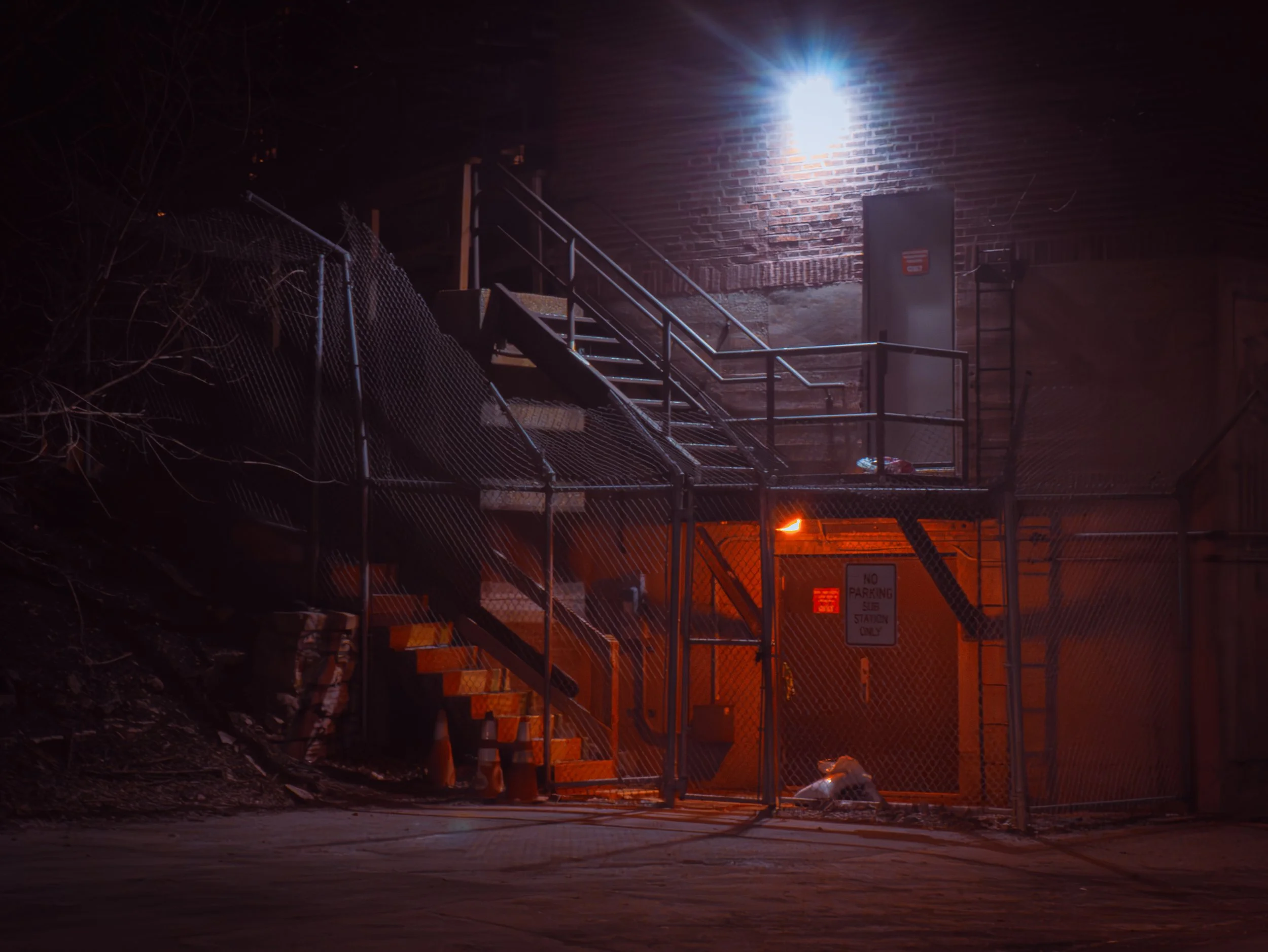 Nighttime scene of a fenced-off construction area with stairs, a brick wall, and bright white light shining from above.