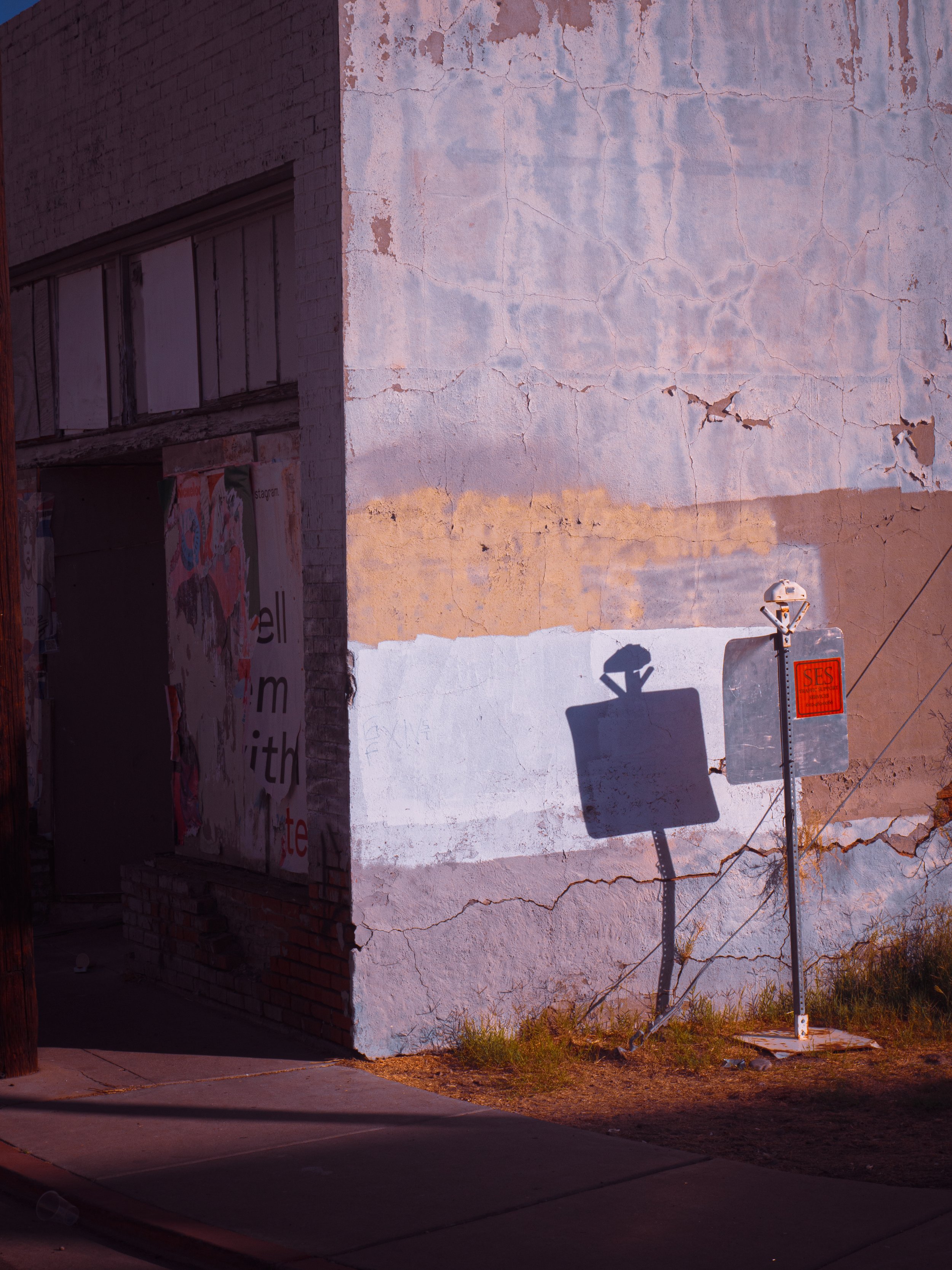 Sunset lighting street scene with an old, cracked, painted wall, a shadow of a streetlamp, and a fallen signpost leaning against the wall.