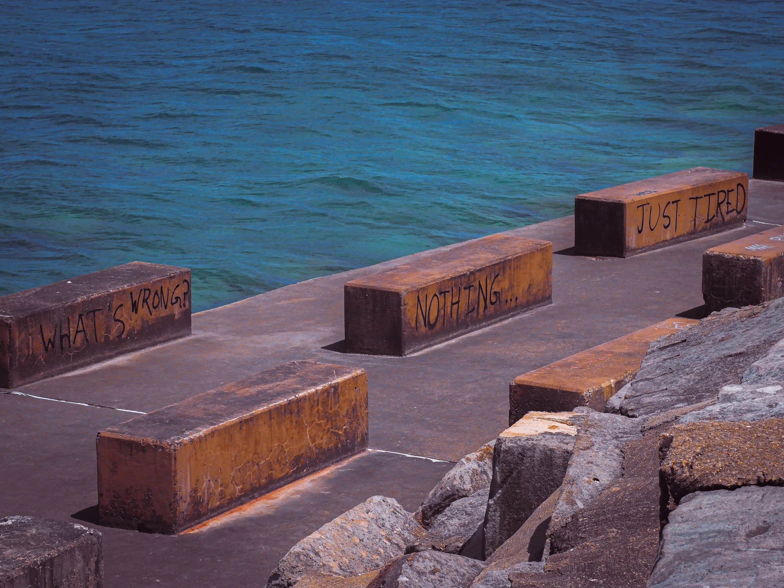 Sea wall with rocks and concrete blocks, some with graffiti, next to blue water.